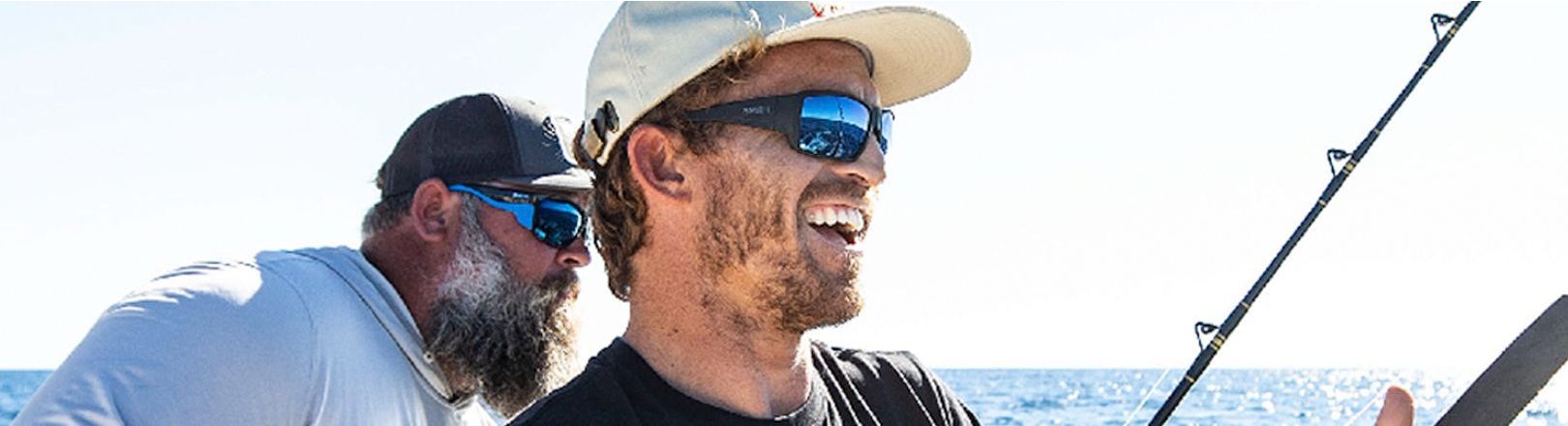 Two men wearing sunglasses and caps smile while fishing on open water. The man in front holds a rod with upward-facing guides, black wrappings, and silver-colored guide frames.