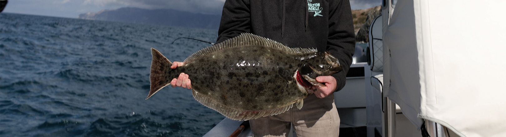 Jason Hower holding a large halibut on a fishing boat; background shows open ocean with distant coastline under cloudy skies.
