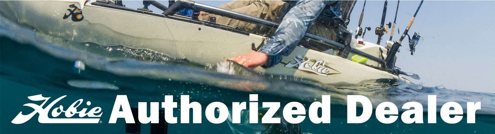 Angler in a tan Hobie kayak reaches into the water beside mounted fishing rods, with “Hobie Authorized Dealer” text in large white letters across the lower part of the image.