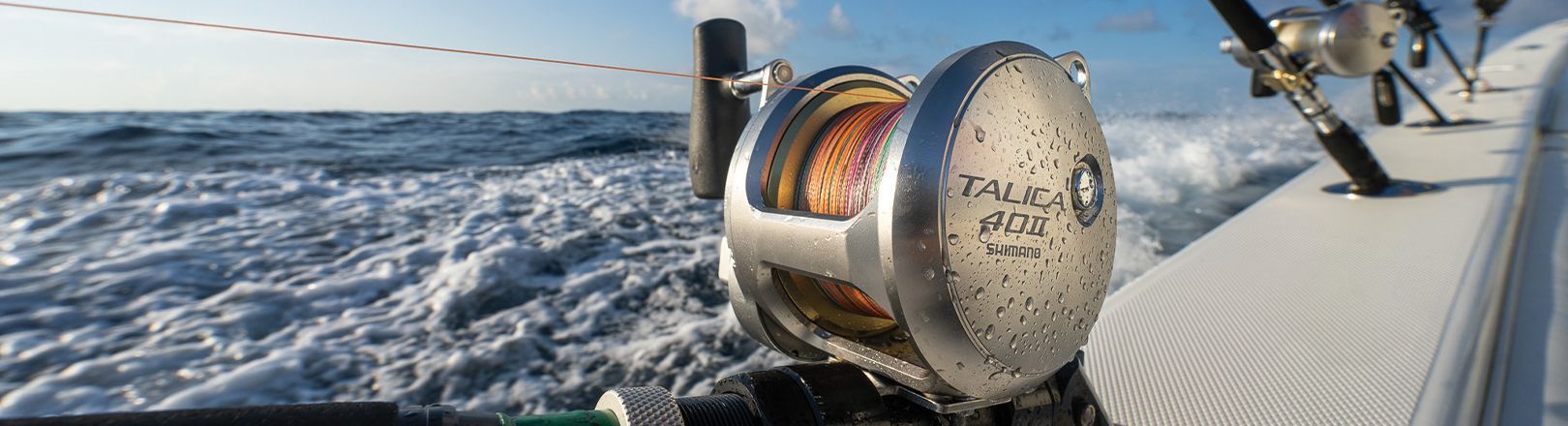 Close-up of a Shimano Talica 40II fishing reel mounted on a boat rod holder. Background shows ocean waves and multiple fishing rods along the boat's rail.