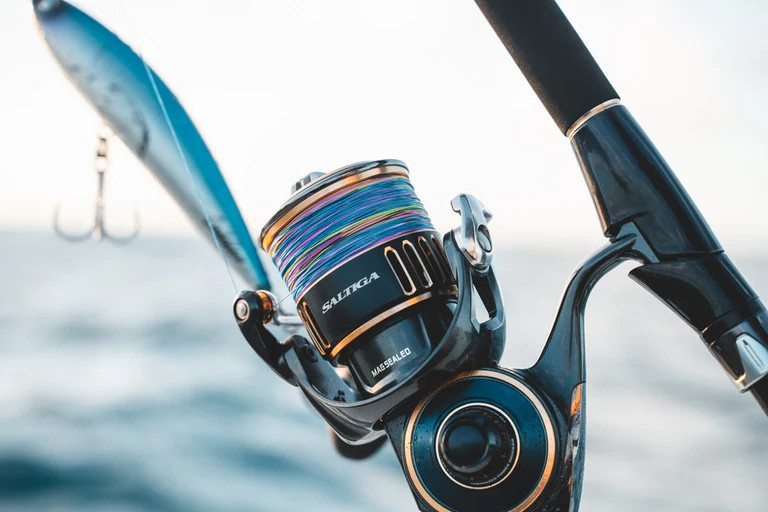 Blue and gold reel spooled with multicolor line on black rod with blue and white lure attached. Blue water and sky in background.