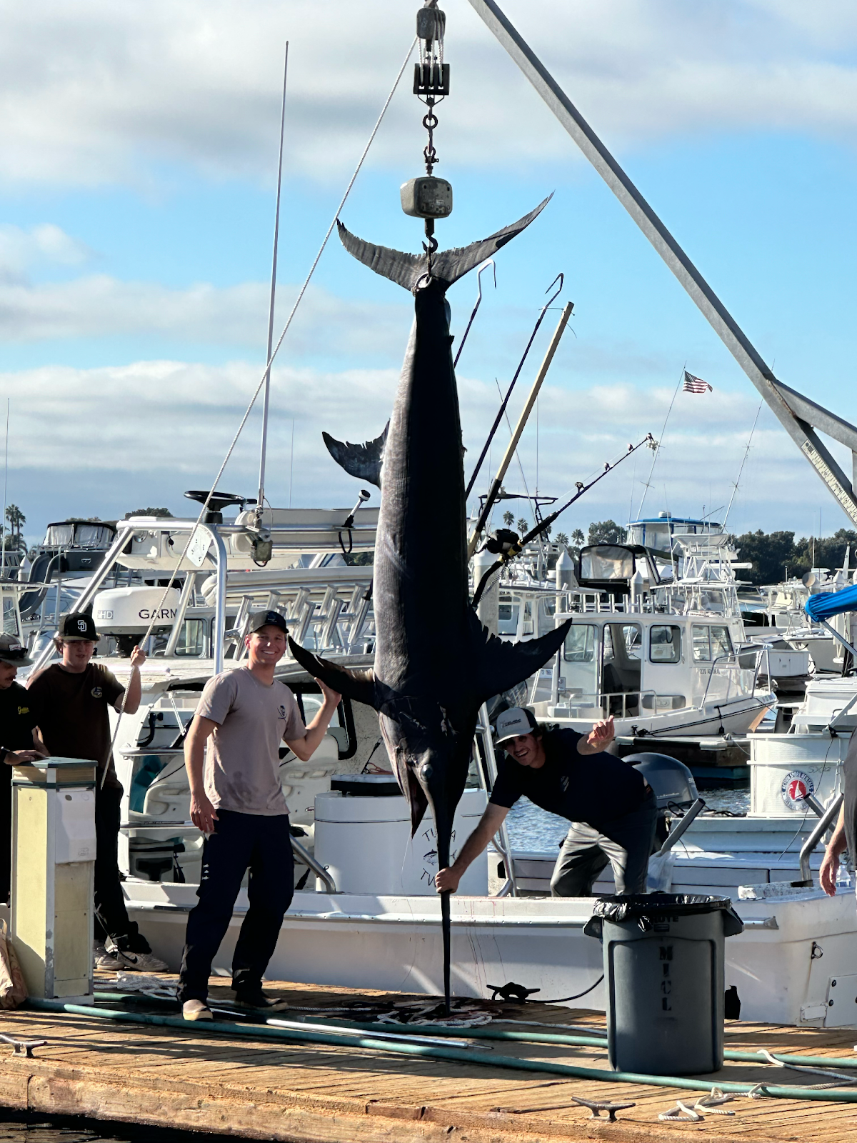 Andy Farmer, Ethan Hougie, and Josh Billauer pose next to their caught 492-pound fish Marlin. The fish is hanging from a crane and the men are smiling broadly.