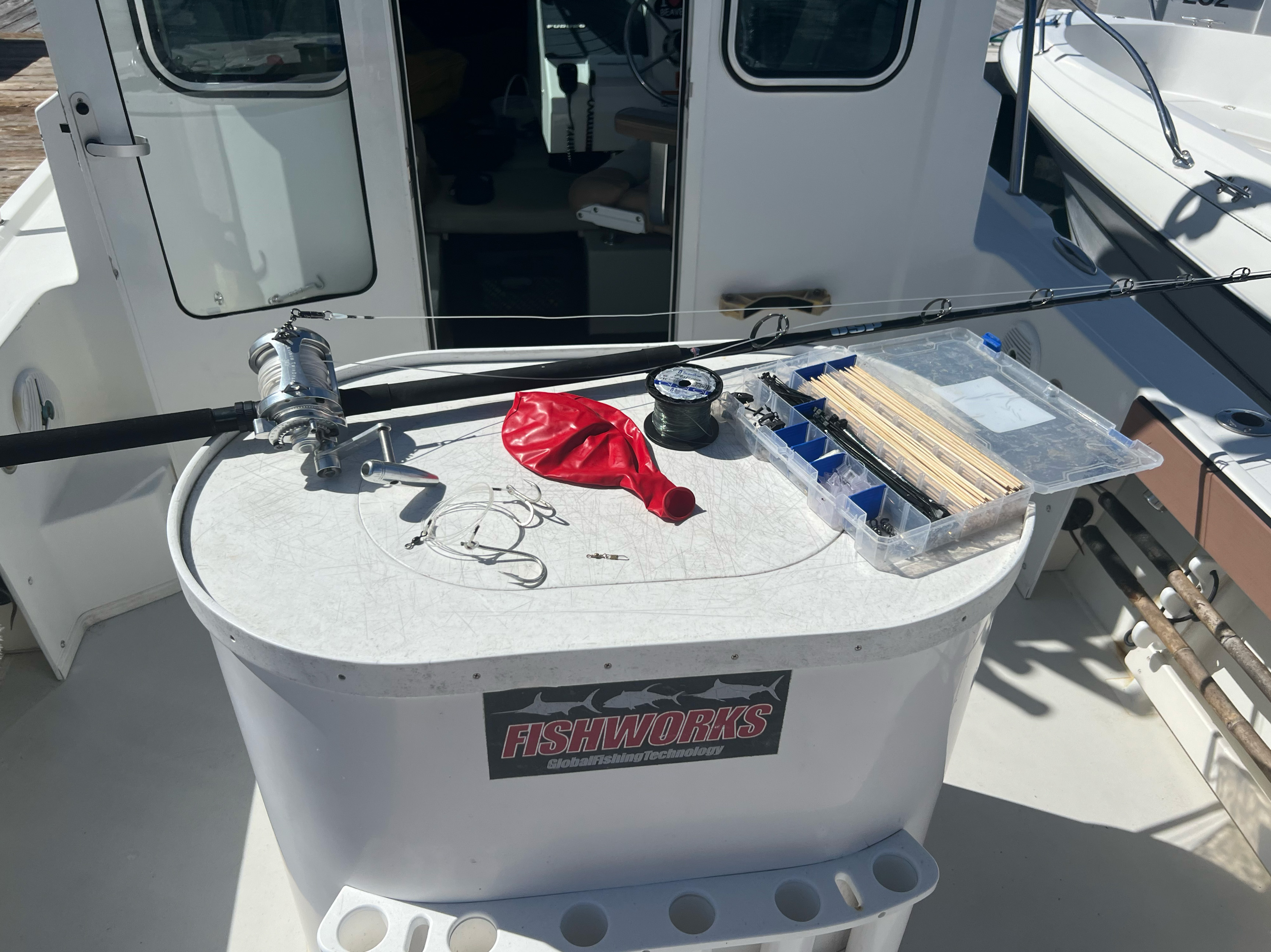 A black rod with silver color reel, hooks rigged onto leader, large red uninflated balloon, spool of dark fishing line, and an open tackle box with crimps, zip ties and wooden sticks laid on a prep table.