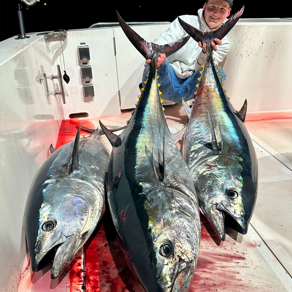 Kyle Farmer kneels on a boat deck holding tails of two of the three large, freshly caught bluefin tuna. The fish have shiny bodies with a dark top and with yellow finlets, and blood is visible on the white deck.