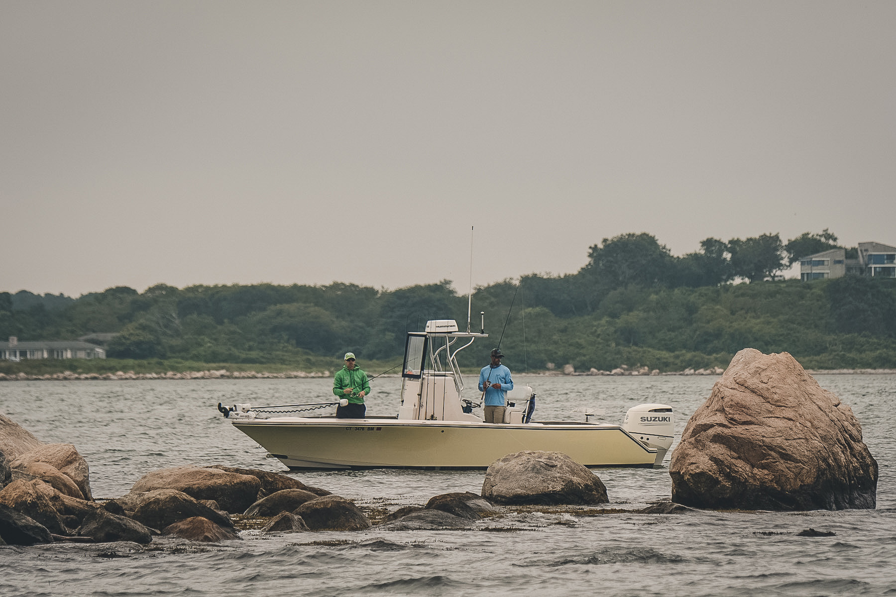 Two people standing on a small boat near a rocky shoreline with a forested hill in the background under gray skies.