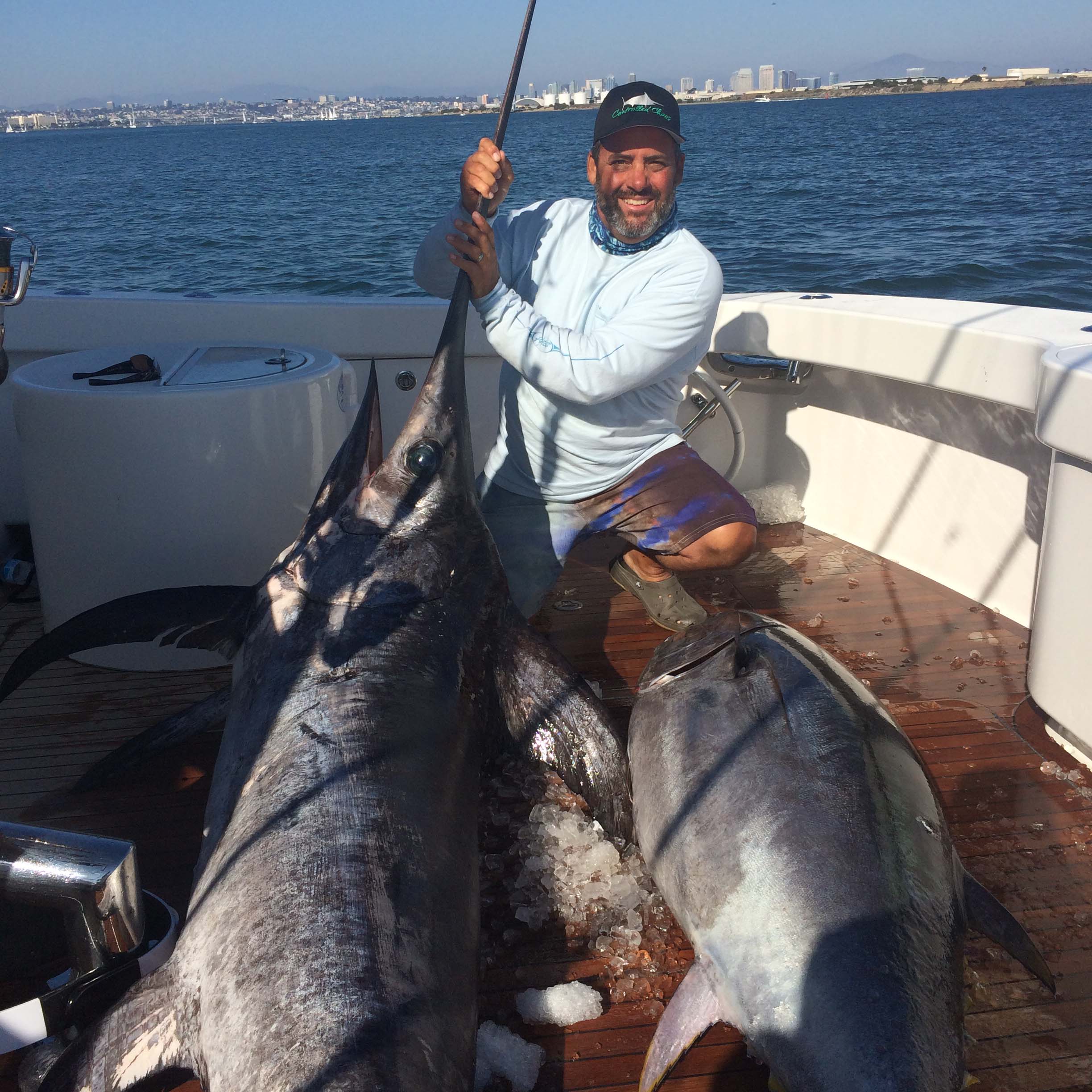 A man on a boat deck holds the bill of a large marlin lying on ice beside another big tuna. He smiles toward the camera with the ocean and city skyline visible in the background.