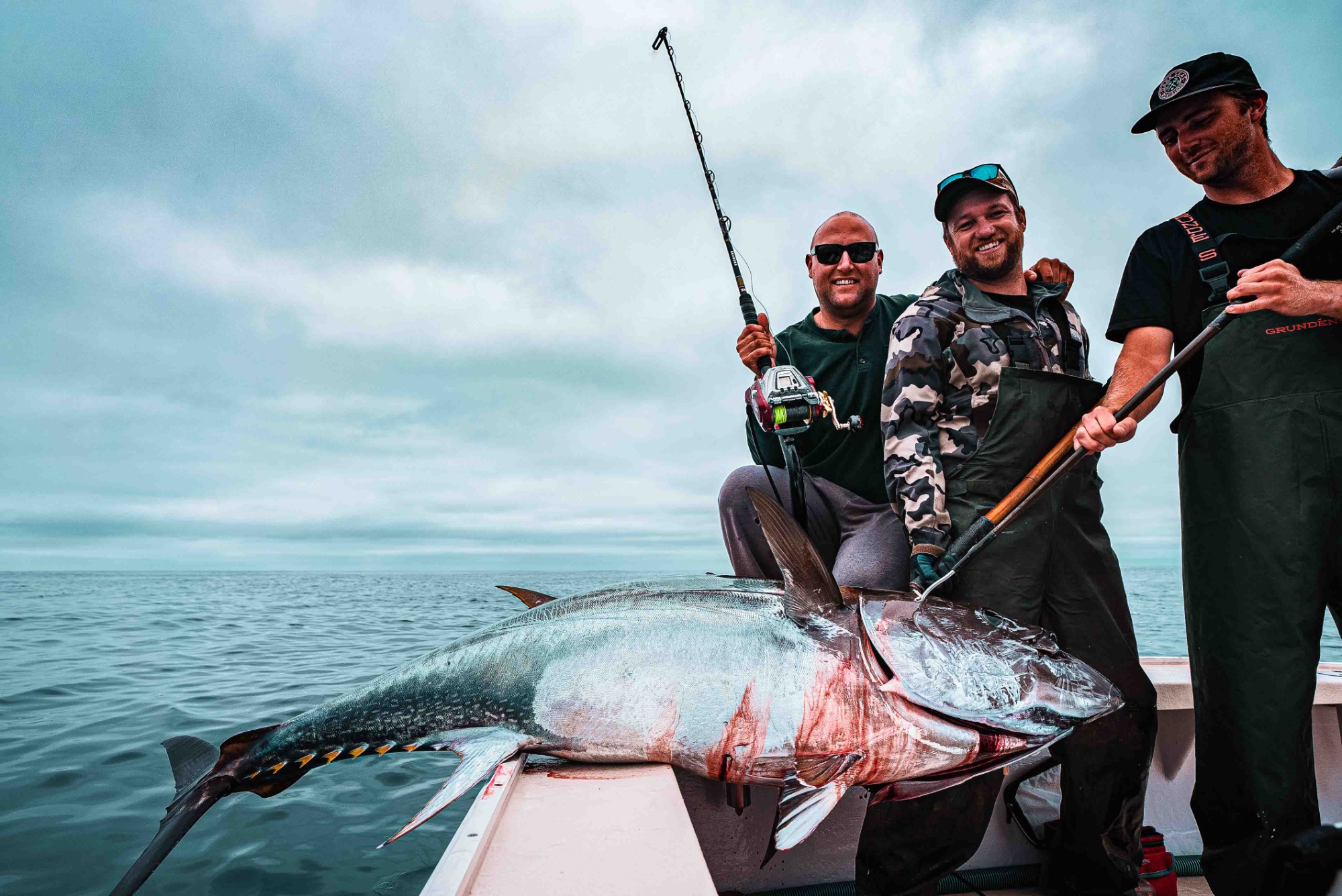 Three men on a boat pose with a giant tuna and a Daiwa electric reel. One holds the rod, another supports the fish. Background shows overcast sky and ocean.
