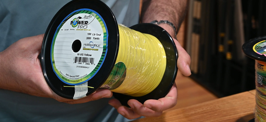 Person holding a large spool of PowerPro Hollow-Ace braided line labeled &ldquo;100 lb Test, 3000 Yards, Hi-Vis Yellow.&rdquo; The spool is filled with bright yellow fishing line and has a black rim.