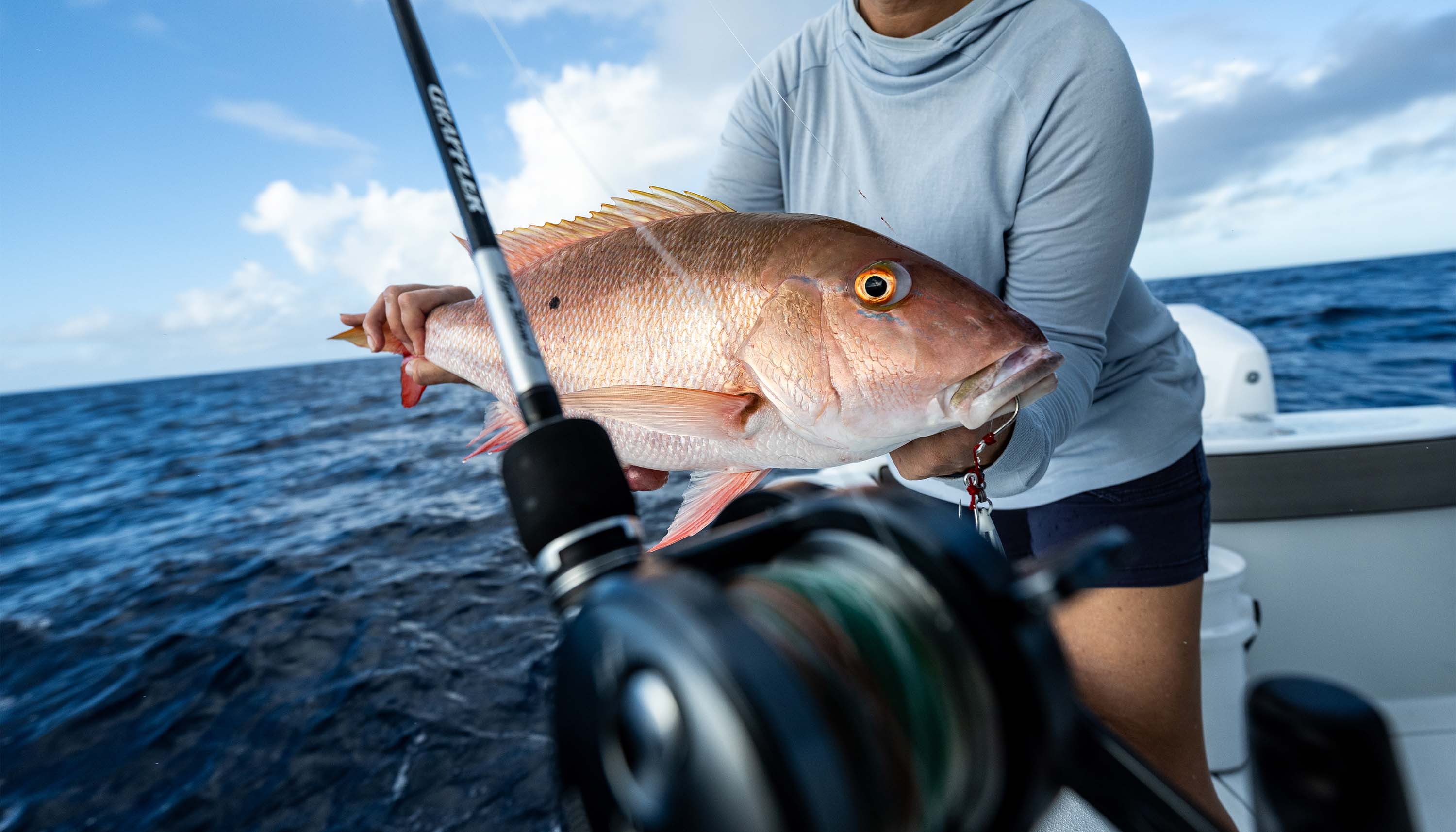 Person on a boat holding a large pinkish-red fish over open ocean water, with a fishing rod and reel visible in the foreground under a partly cloudy sky.