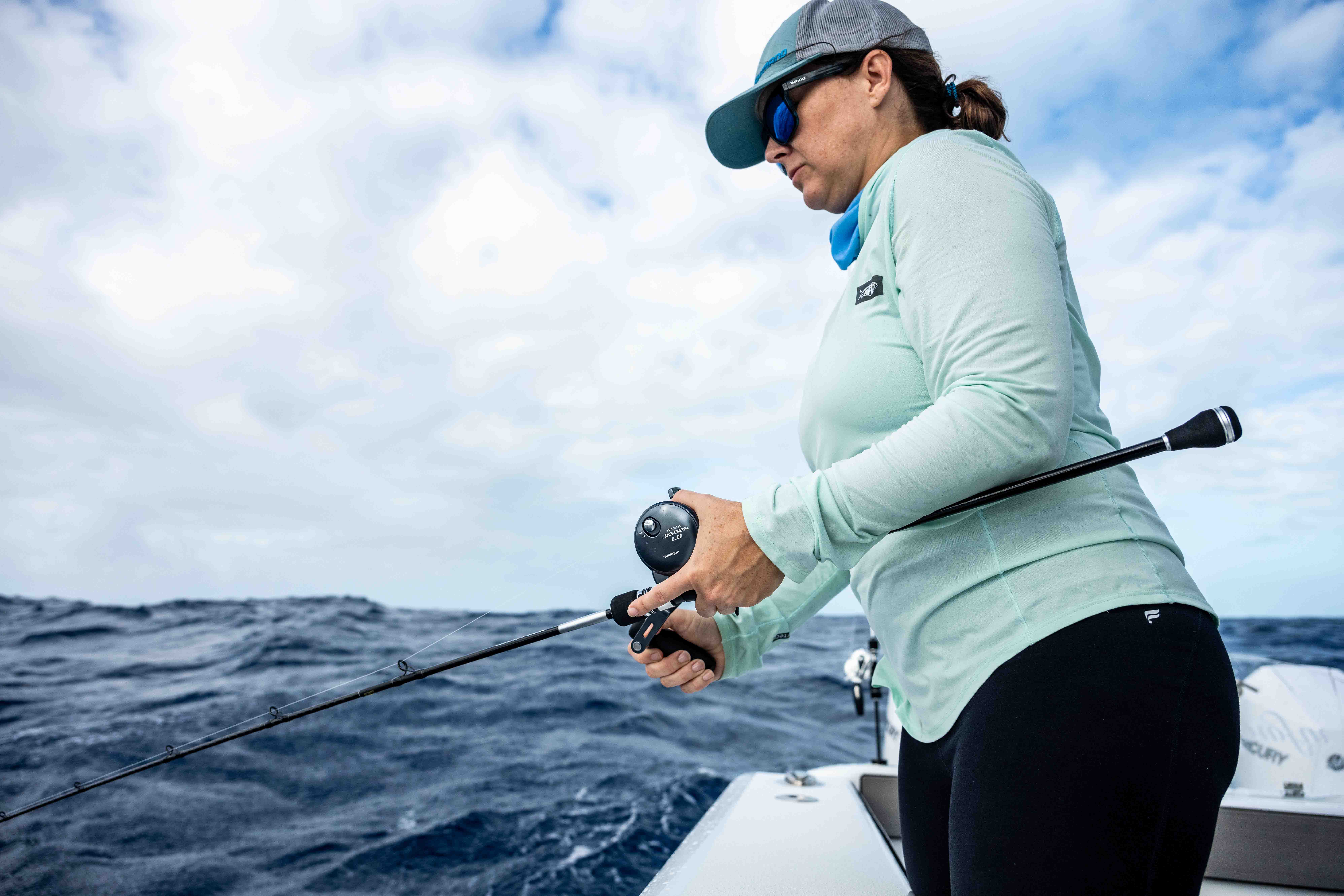 Person standing on a fishing boat, holding a fishing rod over open ocean water while jigging under a cloudy sky.