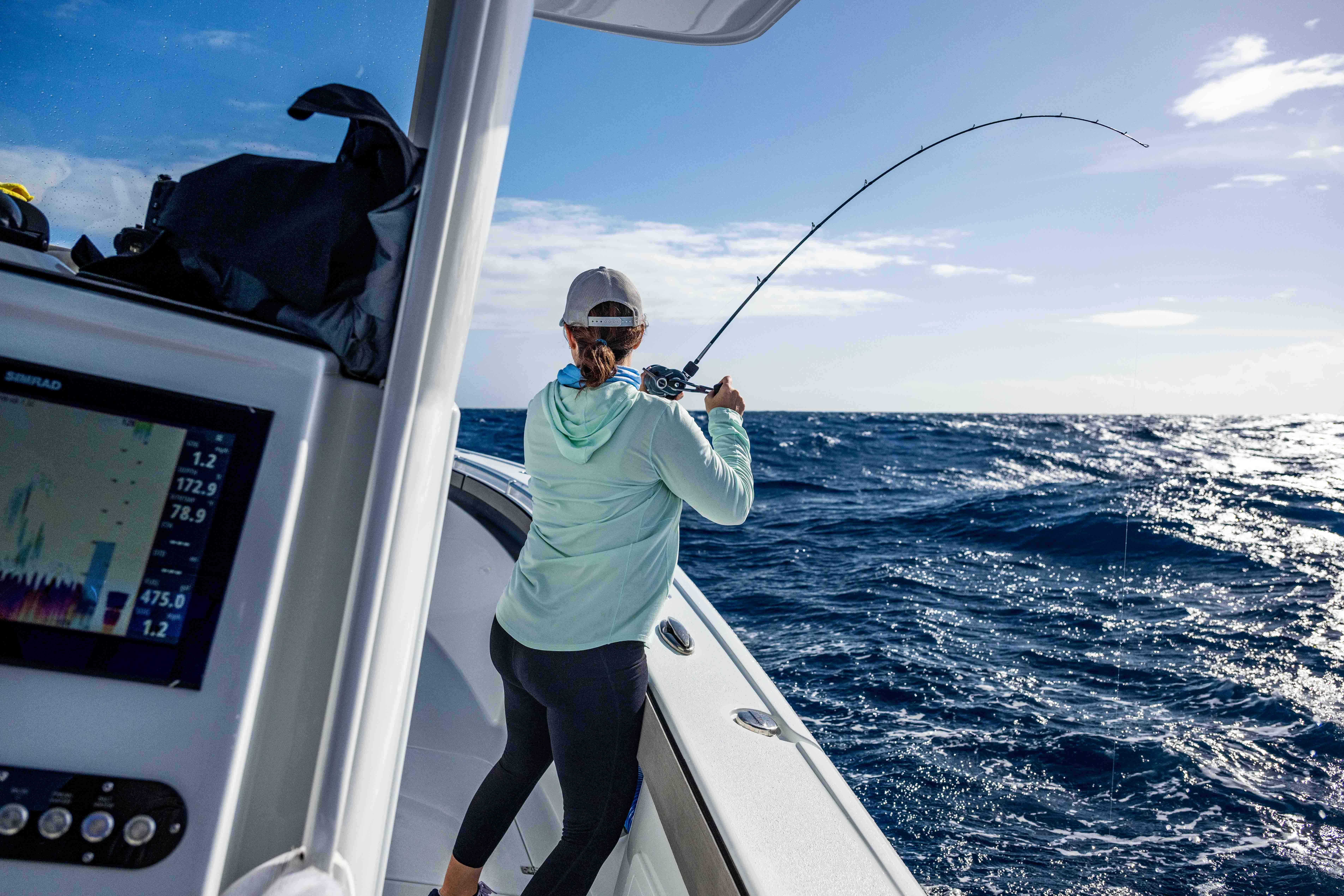 Person standing on a fishing boat fighting a fish, holding a bent fishing rod over rough ocean water under a bright sky.