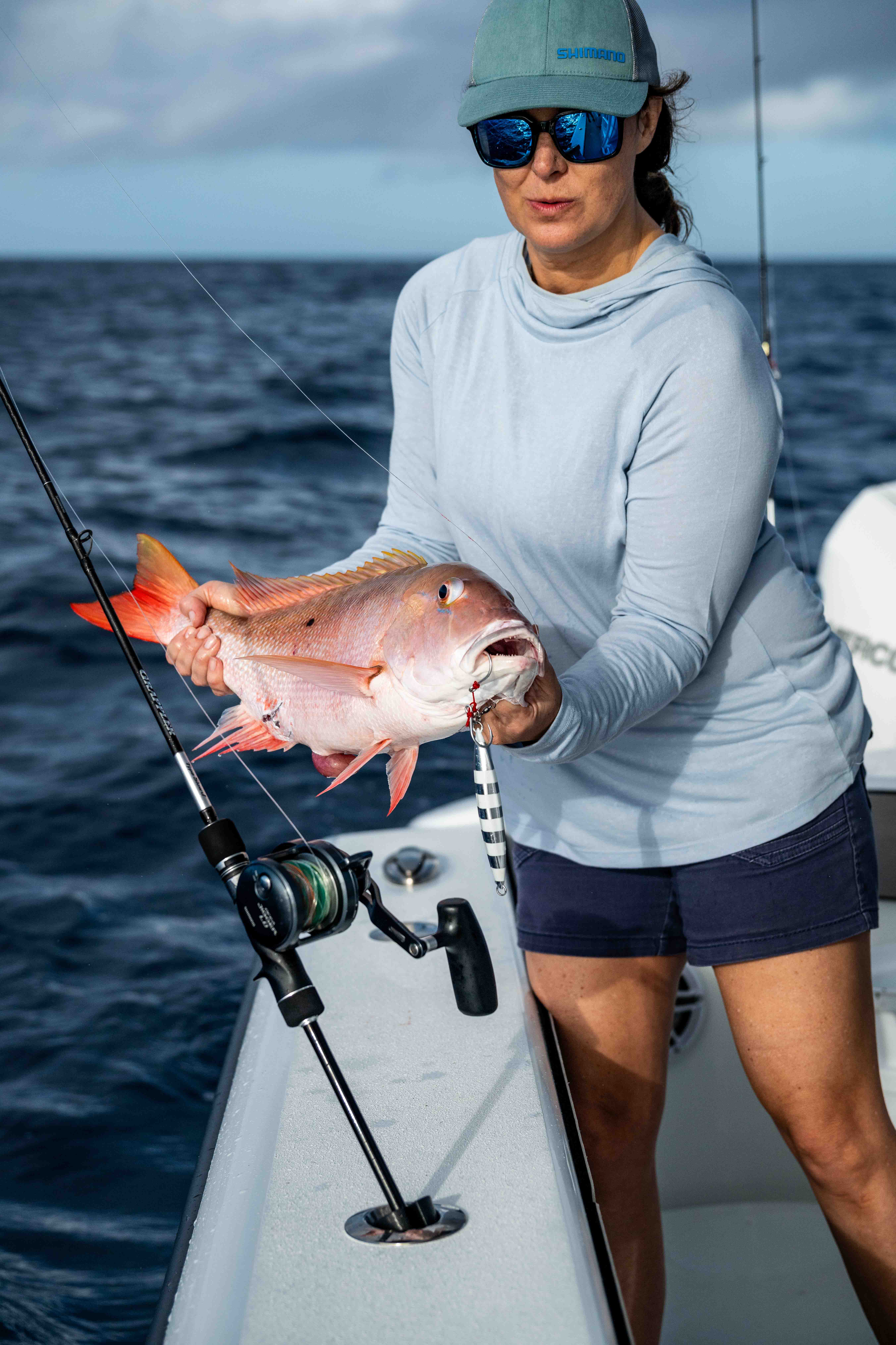 Person standing on a fishing boat holding a freshly caught pink-orange fish beside a fishing rod, with open ocean water in the background.