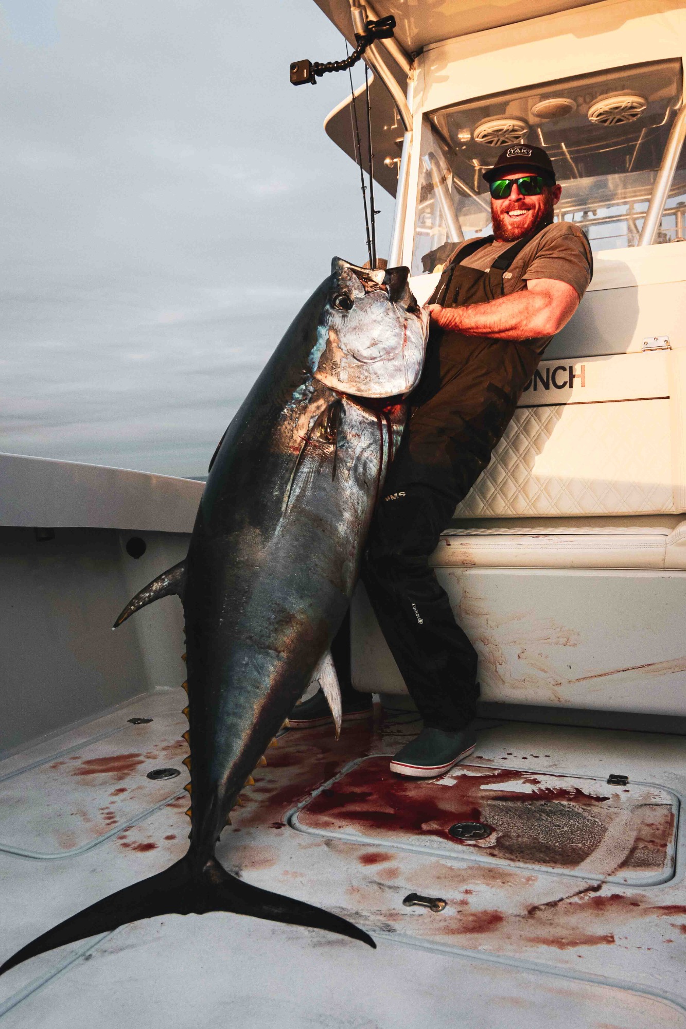 Angler in dark overalls and sunglasses smiles while holding a massive Atlantic bluefin tuna upright on a blood-stained deck of a sportfishing boat.