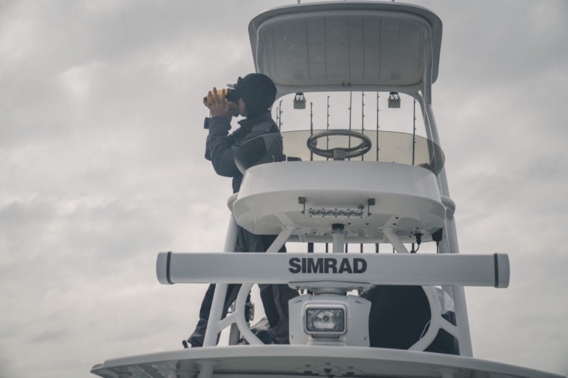 Person standing on the upper deck of a fishing boat using binoculars under a cloudy sky, with fishing rods and navigation equipment visible.
