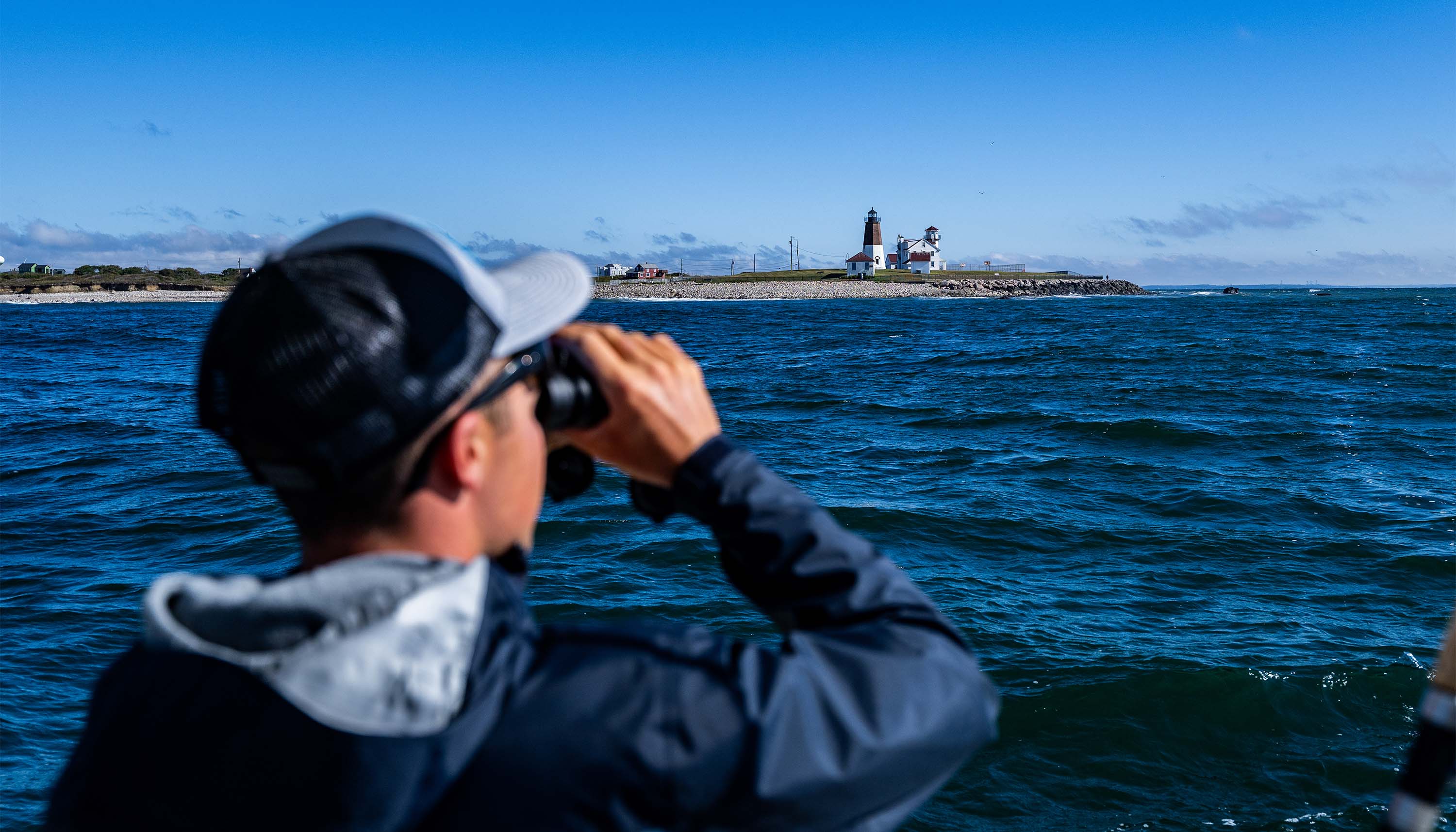 Albie Blog image showing a person using binoculars from a boat while looking toward a lighthouse across the ocean.