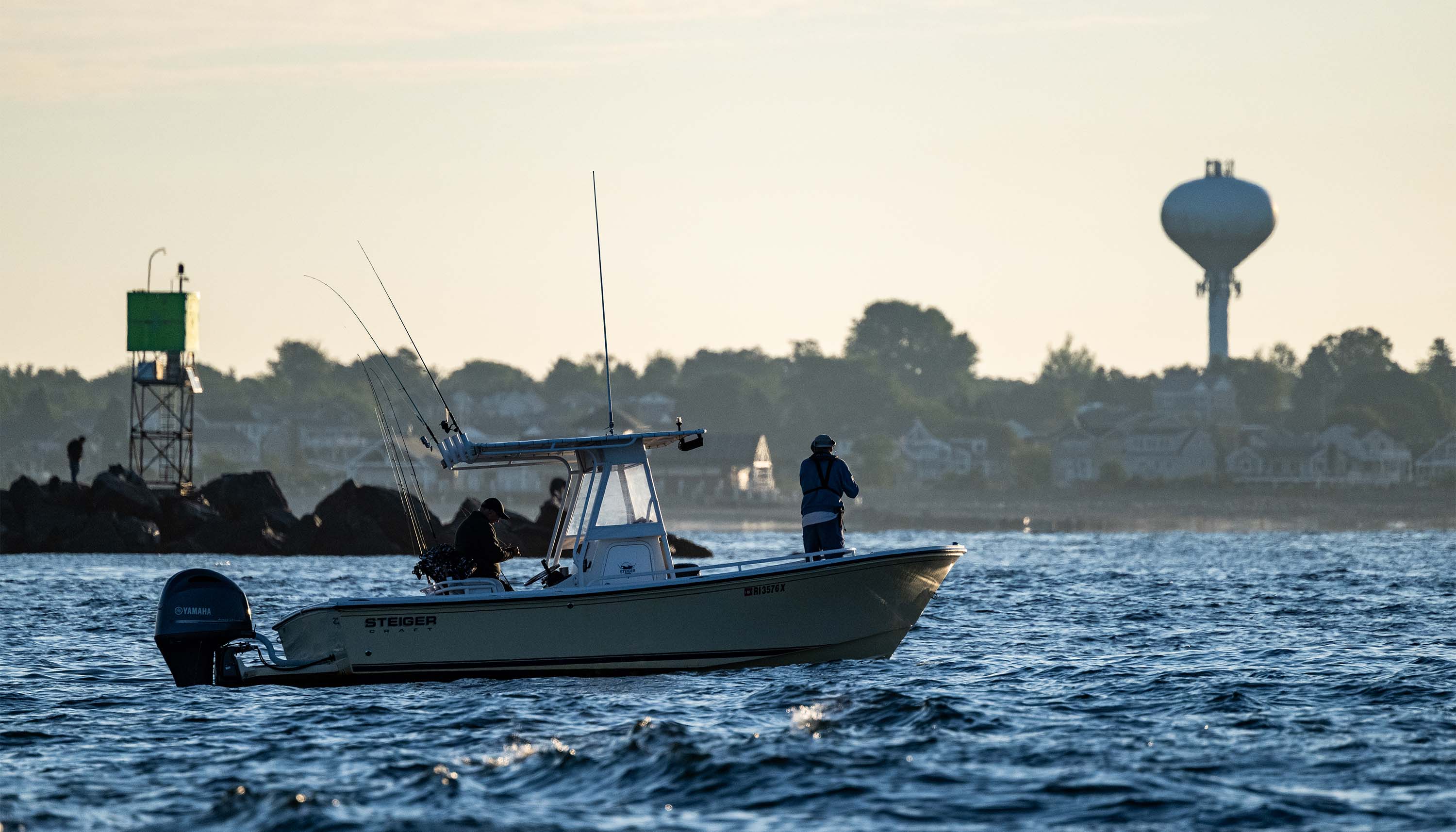 Chasing False Albacore image showing anglers fishing from a boat near a rocky shoreline with a water tower in the distance.