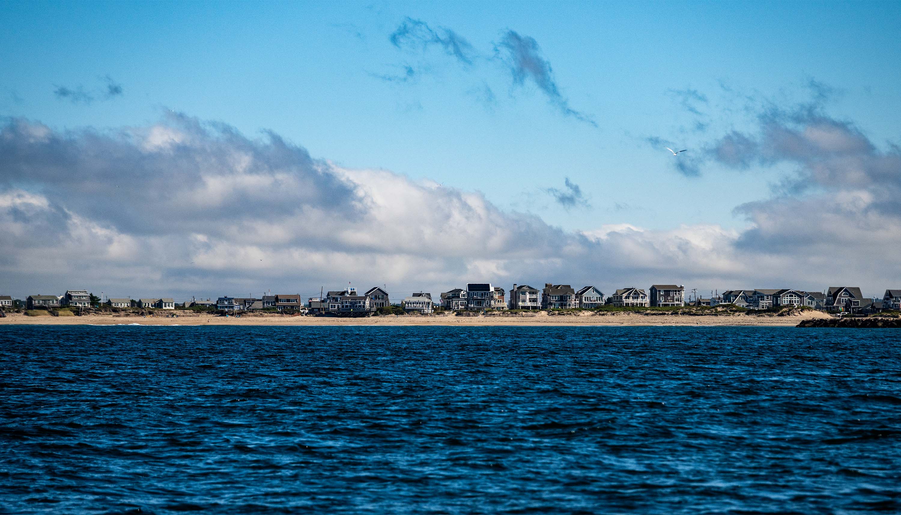 Chasing False Albacore image showing ocean water and a sandy coastline lined with houses under a partly cloudy sky.