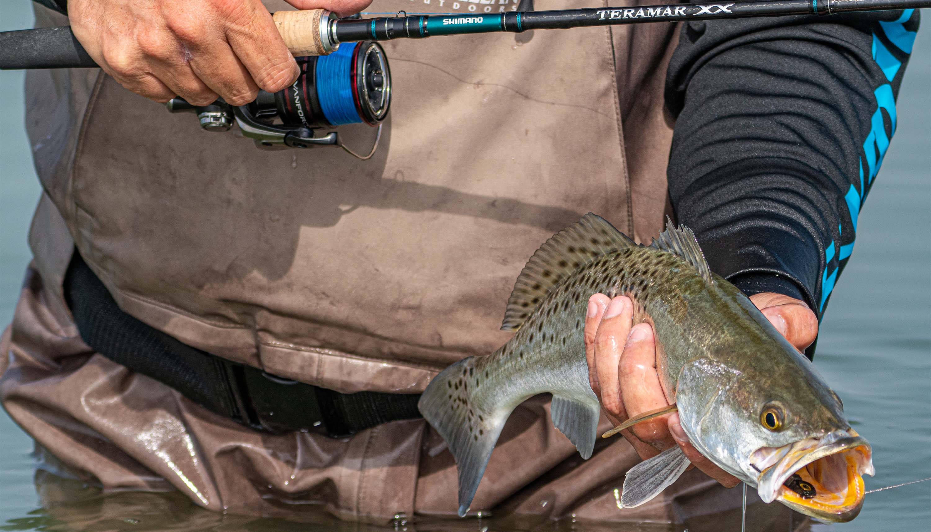 Close-up of an angler standing in shallow water, holding a freshly caught fish beside a spinning rod and reel.