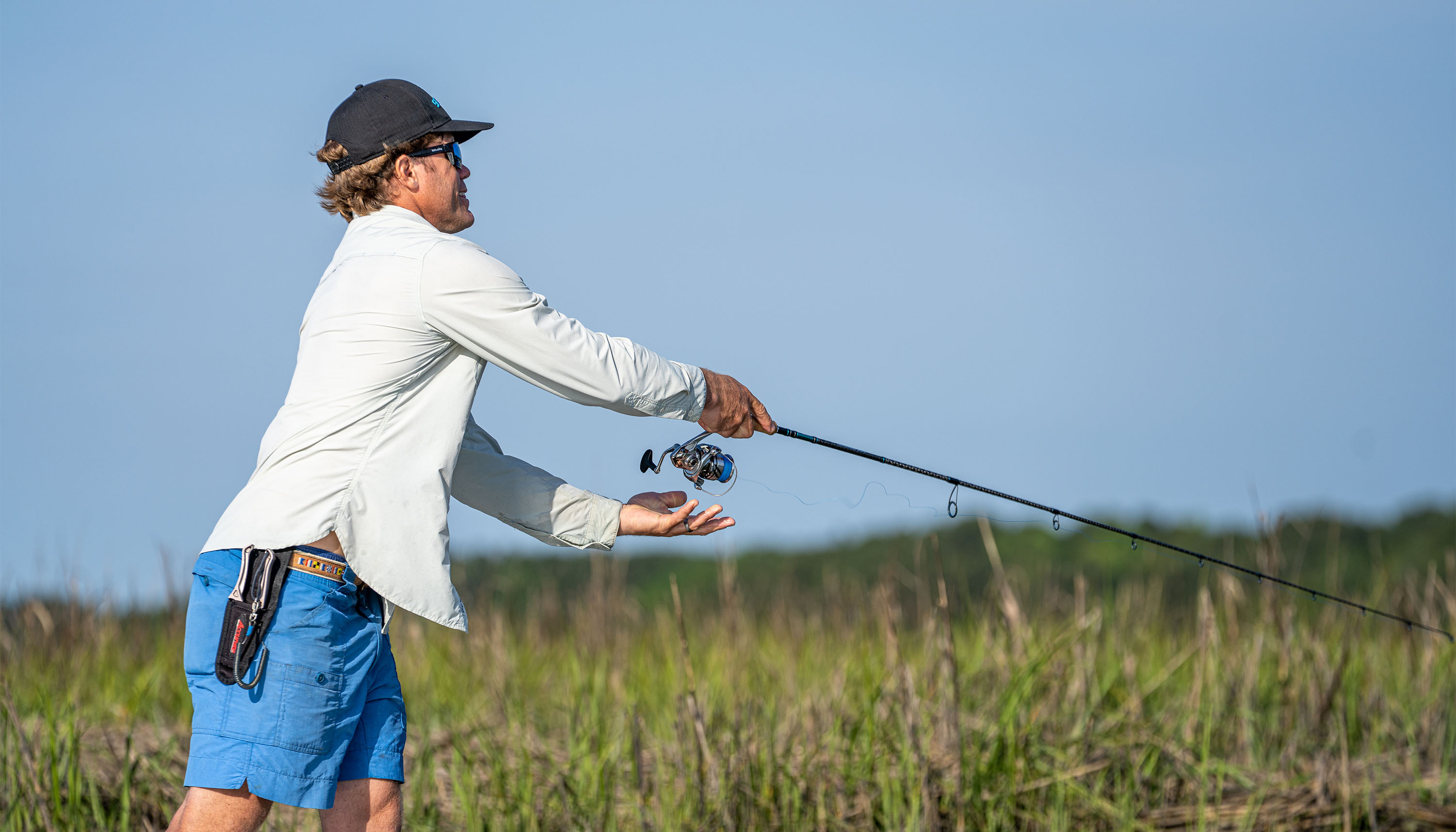 Person wearing outdoor fishing apparel casts a spinning rod while standing beside marsh grass under a clear blue sky.