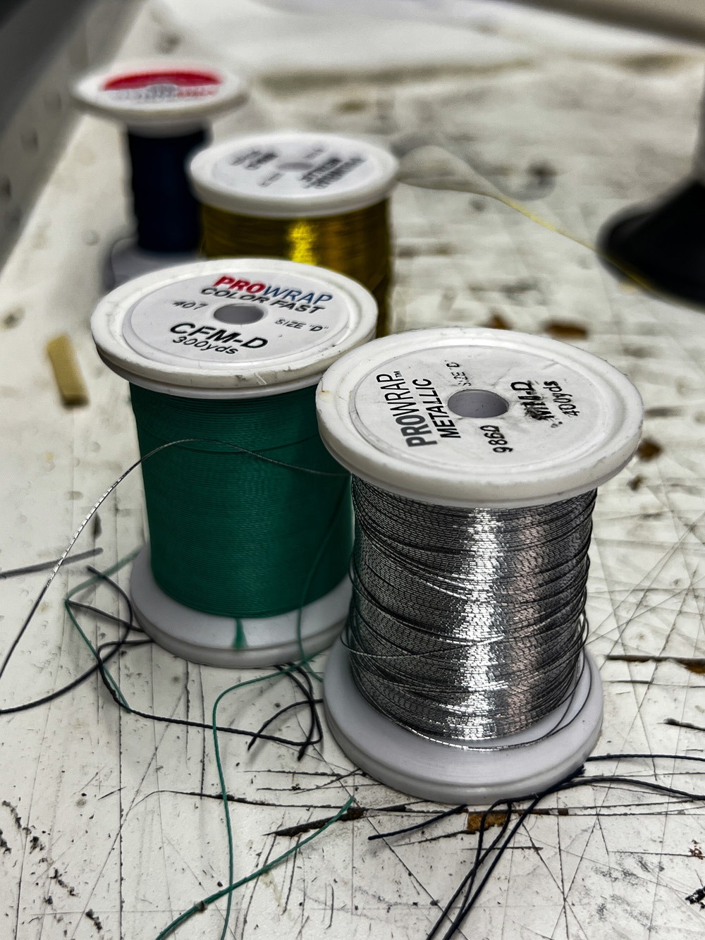 Close-up of spools of thread in silver, green, gold, blue, and red on a scratched white surface.