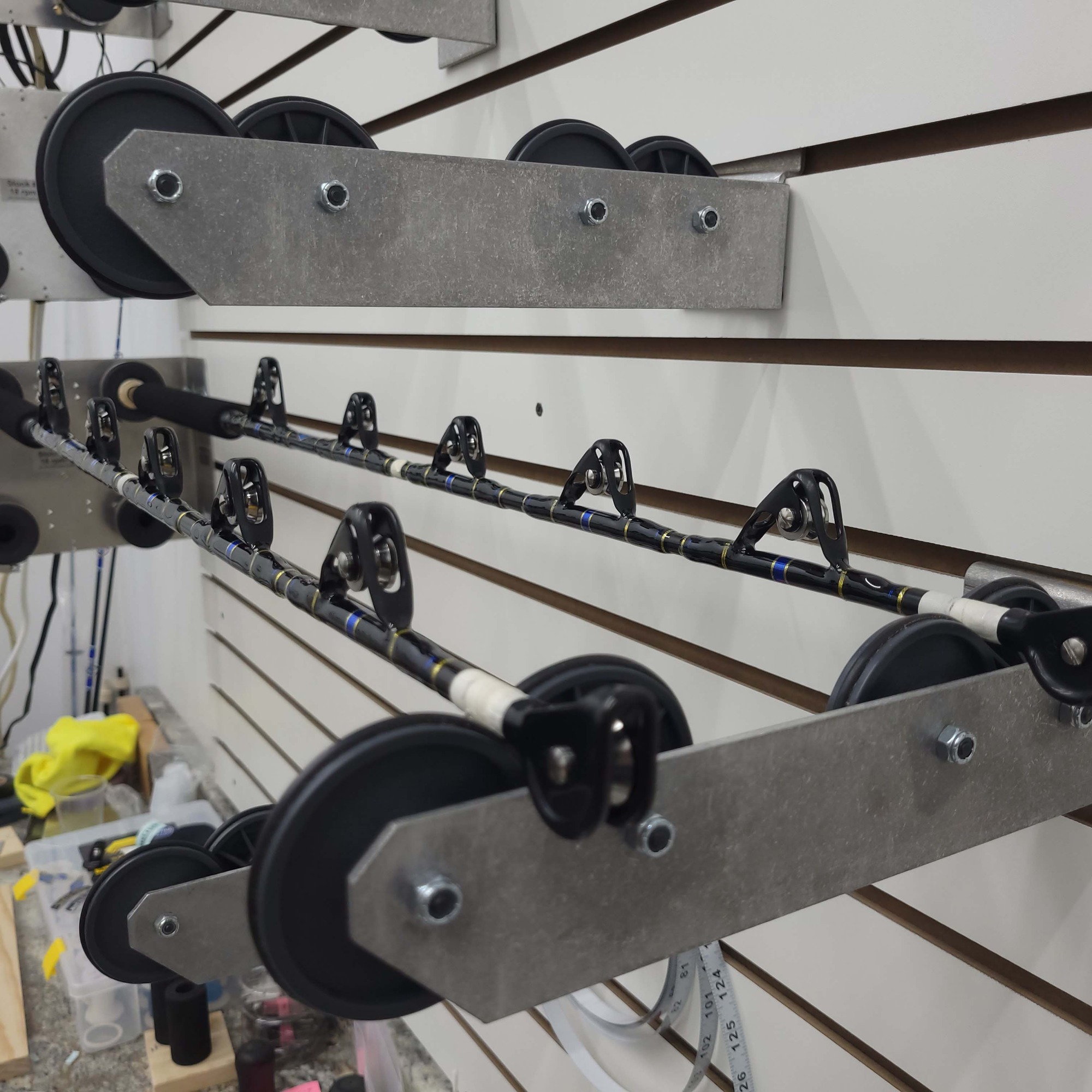 Two custom fishing rods with black and blue guide wraps rest on a horizontal drying rack mounted to a slatwall. Workshop tools and supplies are visible below.