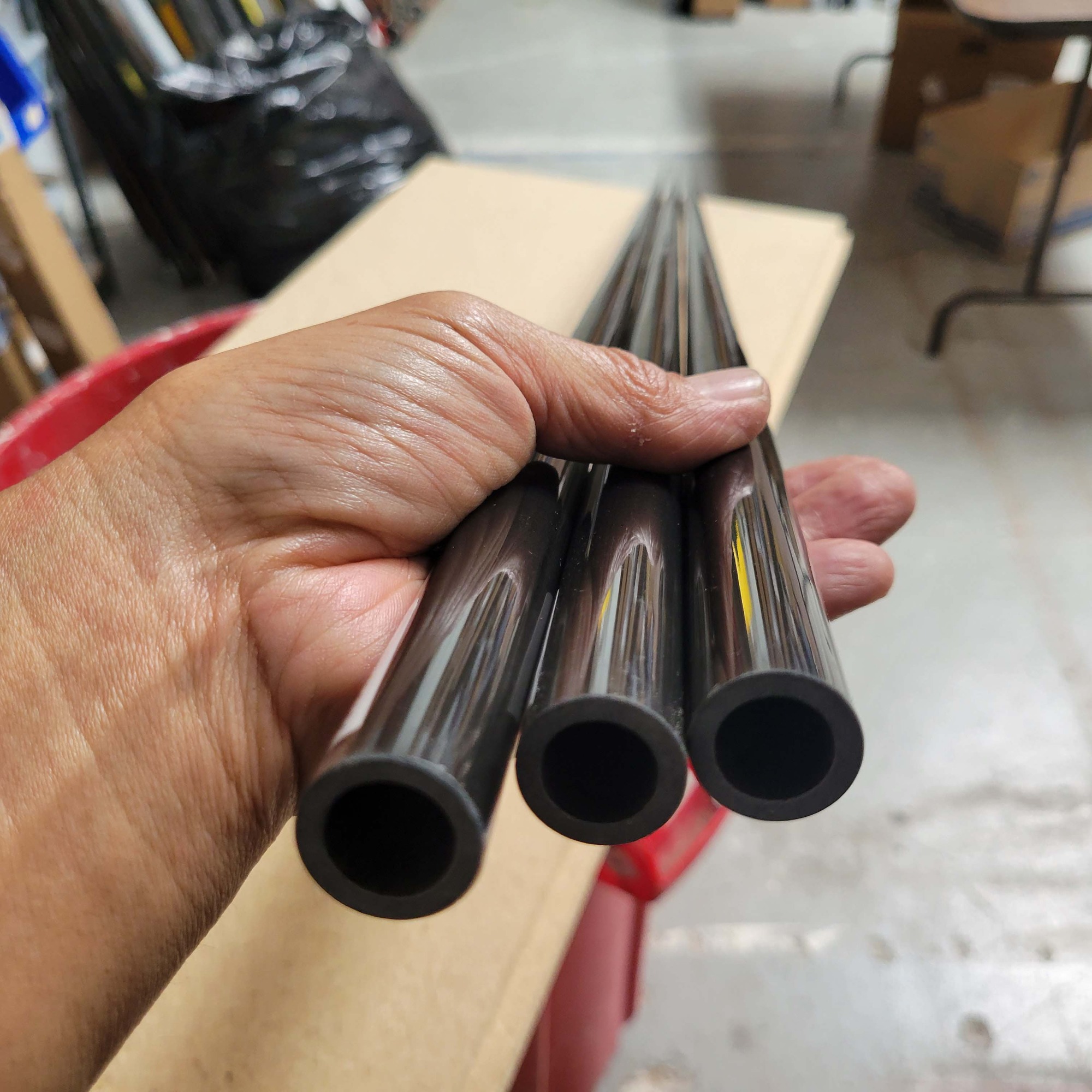 Close-up of a hand holding three glossy black fishing rod blanks, showing their hollow openings. Workshop floor and cardboard surface are visible in the background.