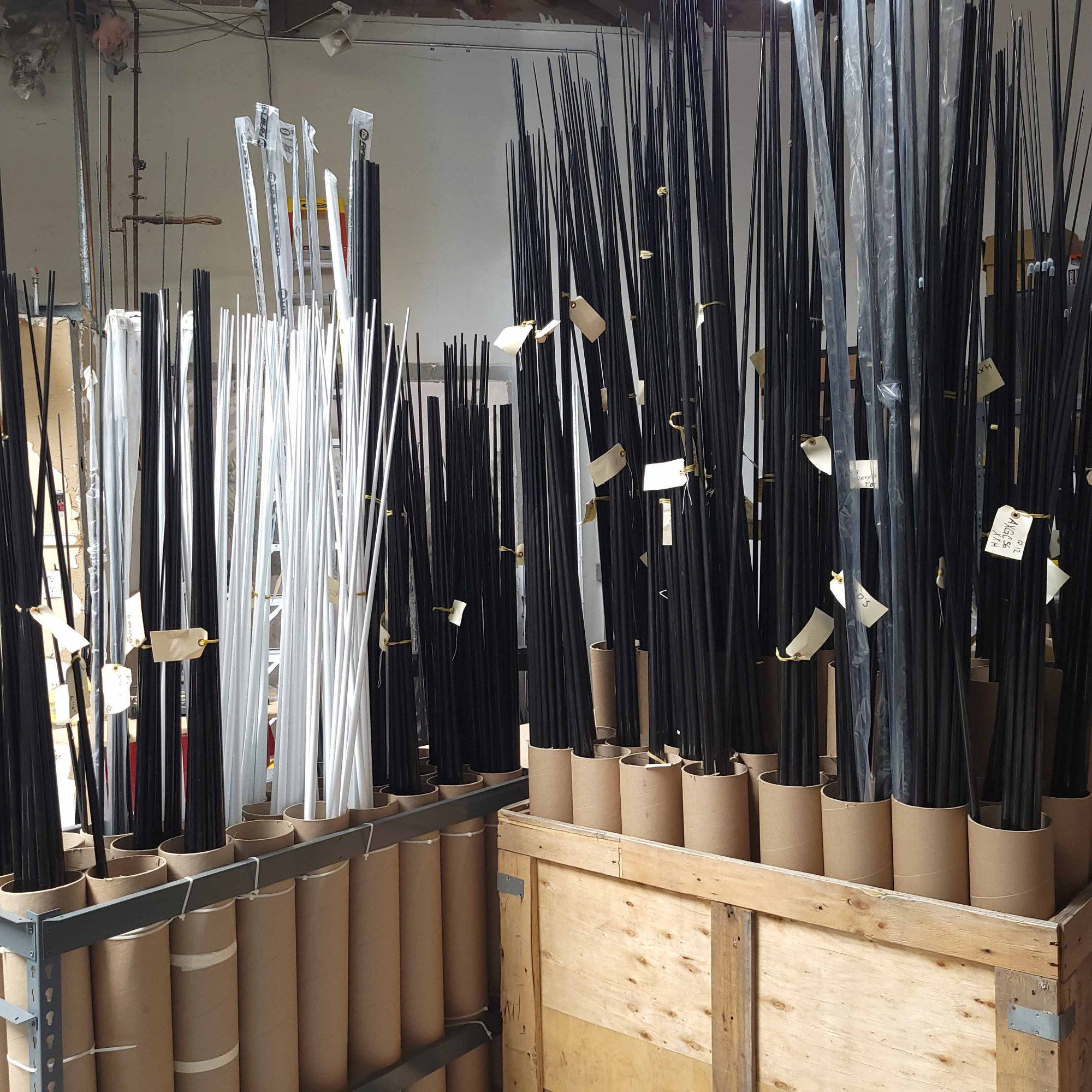 Dozens of black and white fishing rod blanks stand upright in cardboard tubes, organized by size and tagged for inventory in a workshop storage area.