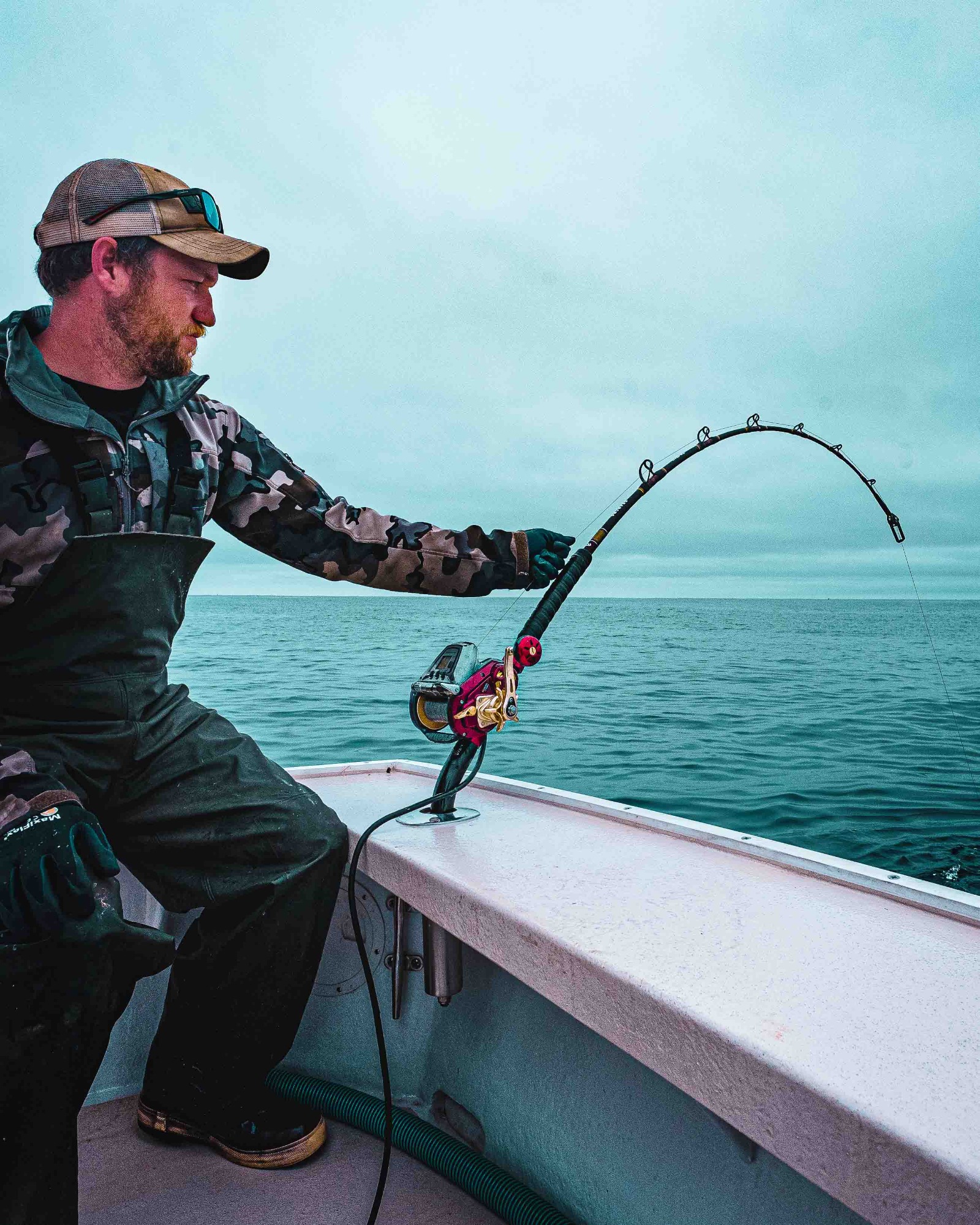 Man seated on a boat in camo outerwear, gloves, and a cap uses a Daiwa electric reel, gripping a bent fishing rod over calm ocean water beneath a cloudy sky.