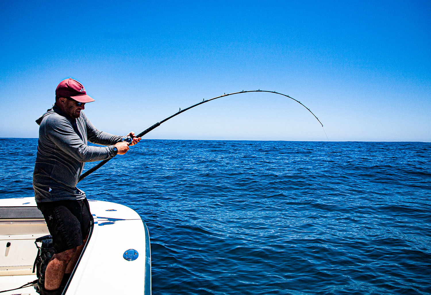 A man on a boat fights a fish with a bent fishing rod, wearing a hoodie and red cap. Background shows open ocean under a clear blue sky.