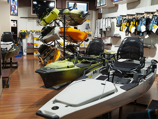 Indoor display of Hobie kayaks in gray, green, yellow, orange, and blue, with two pedal-drive models in front. Wall shelves show paddles, life vests, and accessories arranged on a wooden floor.