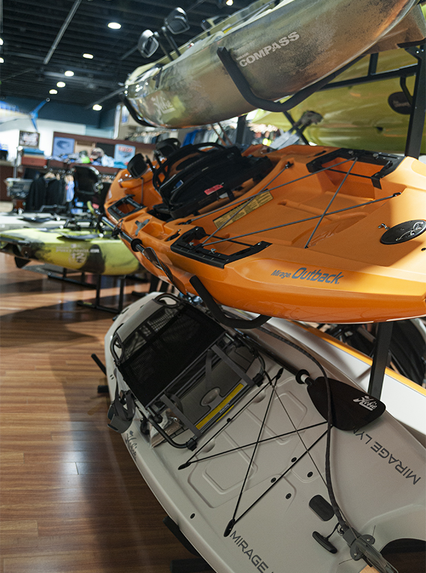 Showroom display of Hobie kayaks stacked on a rack, including green, orange, and white models, with additional kayaks visible in the background on wood flooring.