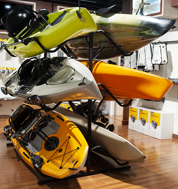 Stacked display of Hobie kayaks in green, gray, orange, and yellow on a black metal rack inside a showroom, with boxed accessories and hanging gear visible along the wall in the background.