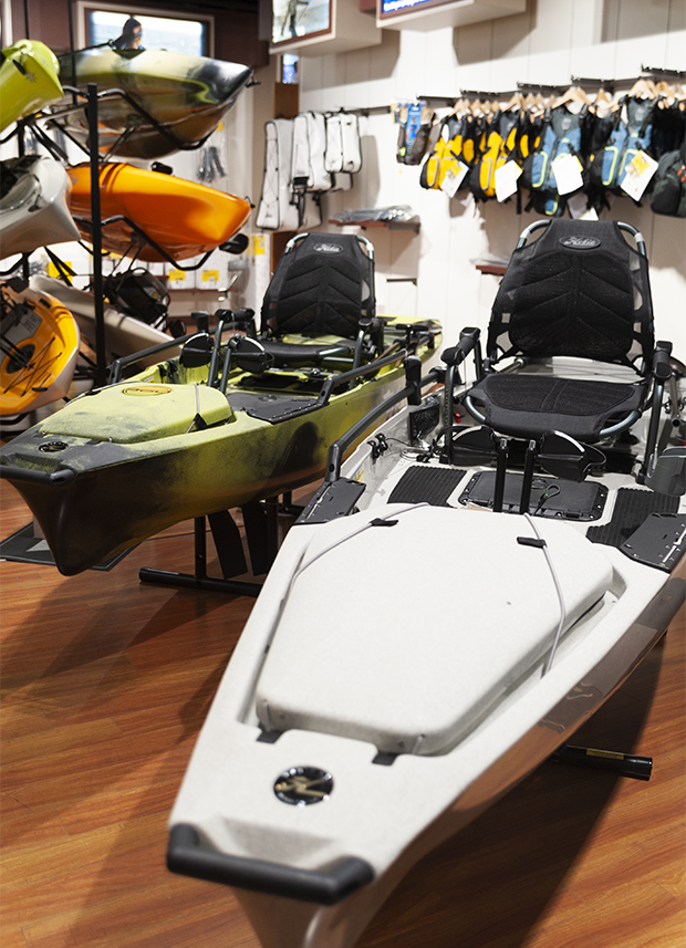 Two Hobie pedal-drive kayaks in gray and green displayed on a wooden floor, with stacked kayaks, paddles, and yellow life vests hanging on the wall in the background inside a retail showroom.
