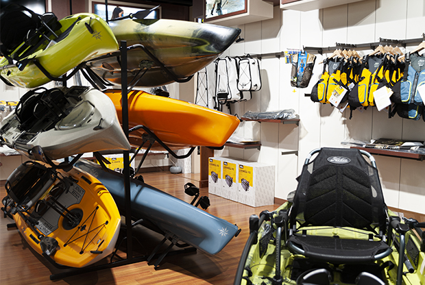 Showroom display of stacked Hobie kayaks in green, yellow, orange, gray, and blue, with paddles, life vests, and accessories hanging on the wall beside boxed products on a wooden floor.