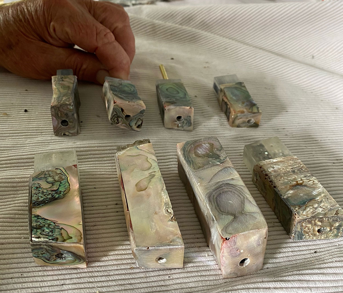 Abalone Shell Inserts Hand near several rectangular abalone shell blocks with iridescent surfaces in beige, green, and pink tones, each drilled through the center and arranged on a light fabric surface.