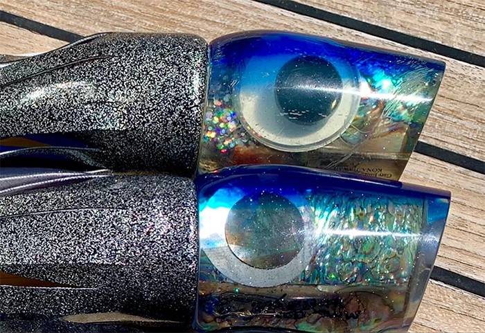 Two Marlin Magic trolling lures with silver glitter skirts and blue-tinted resin heads featuring large reflective eyes and abalone-style inserts, shown side by side on a wooden deck surface.