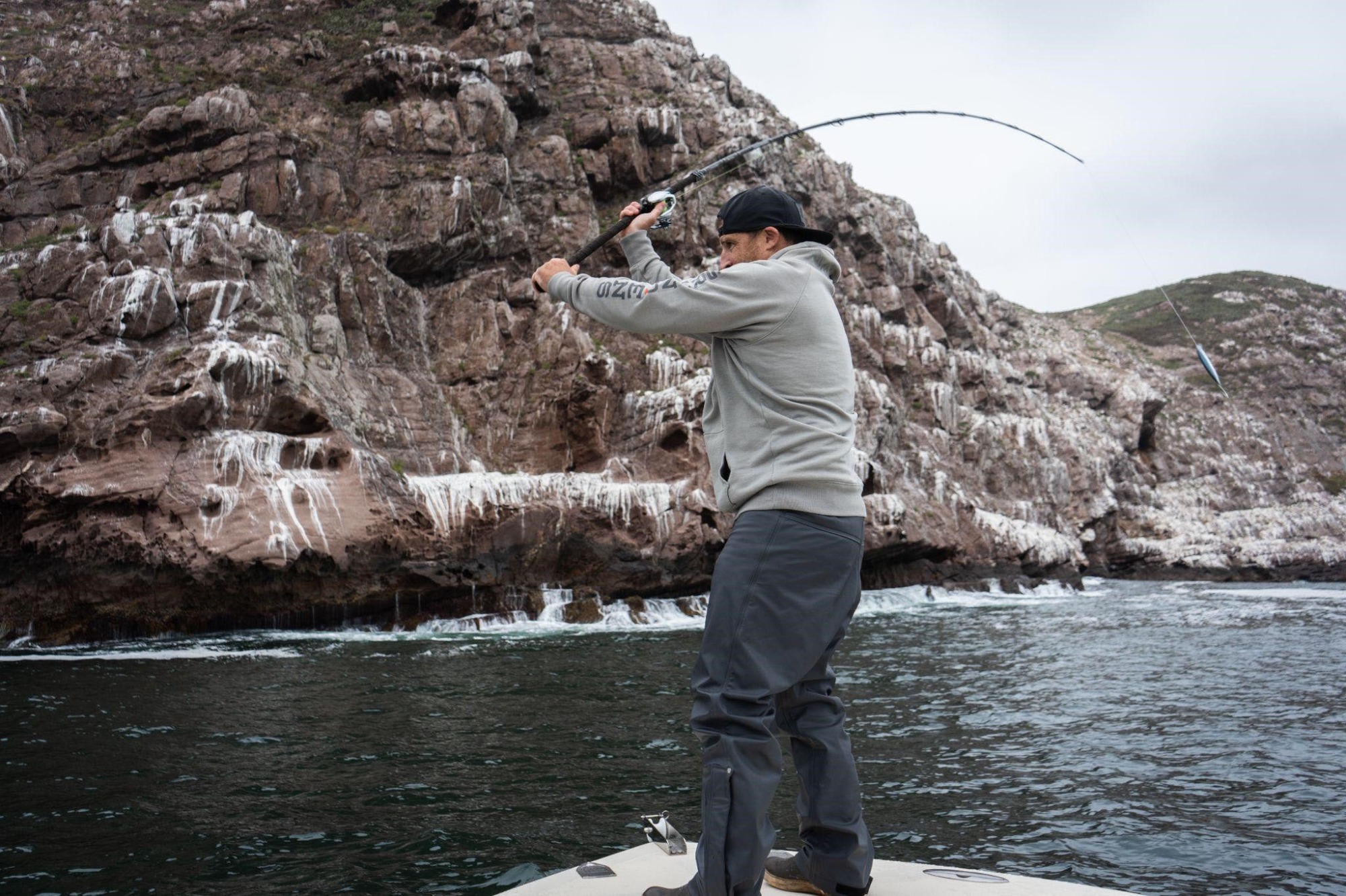 Angler casting a baitcasting reel from a boat near rocky cliffs under an overcast sky.