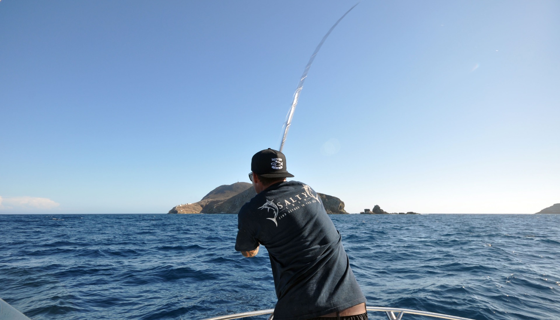 Captain Duane Diego wearing a black cap backwards casts into the ocean. The weather is sunny and the waters are calm.