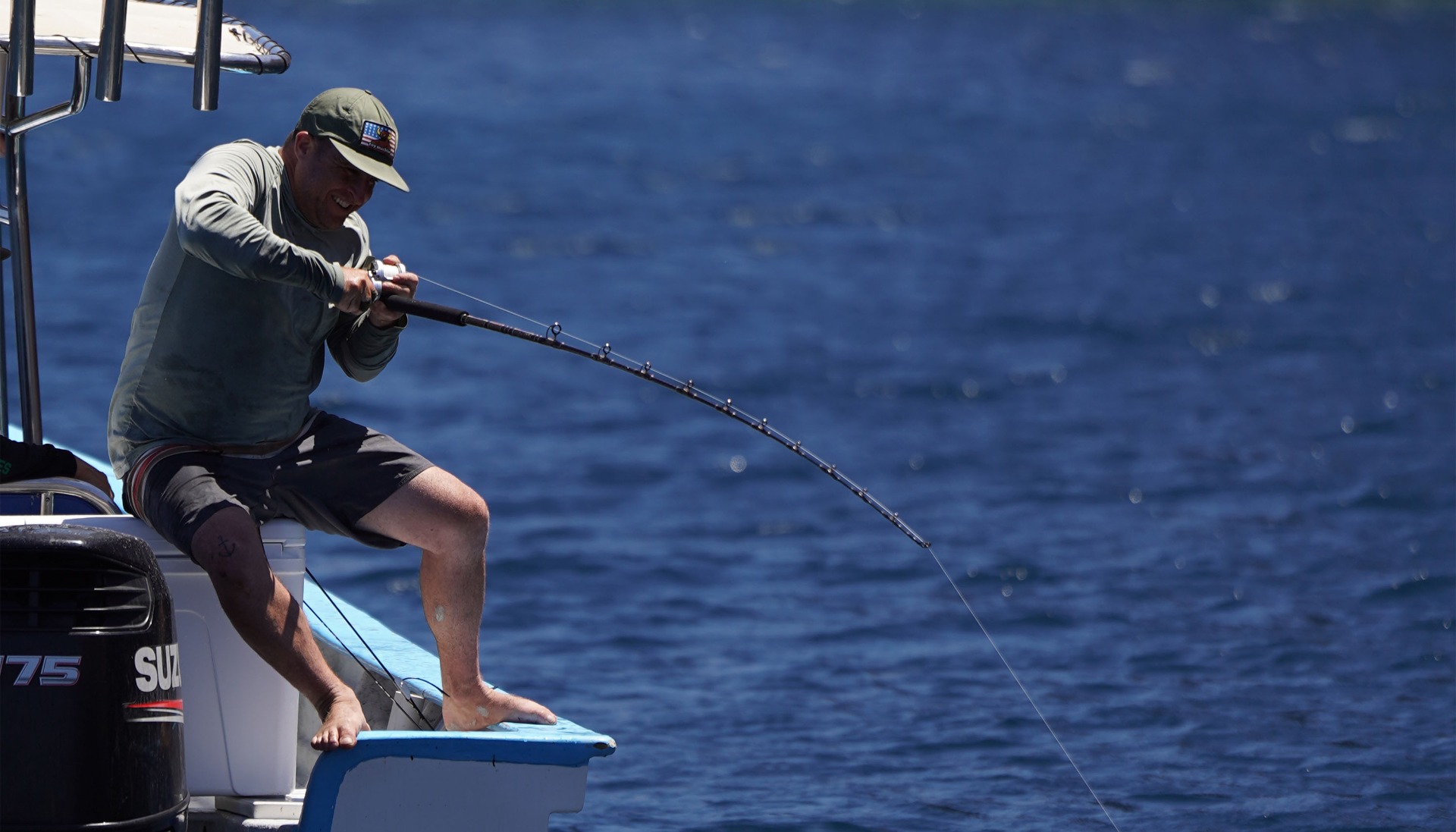 Michel Murciano in green long-sleeve shirt and shorts pushing himself hard against the edge of a boat, gripping a bent fishing rod over deep blue water, indicating a strong catch.