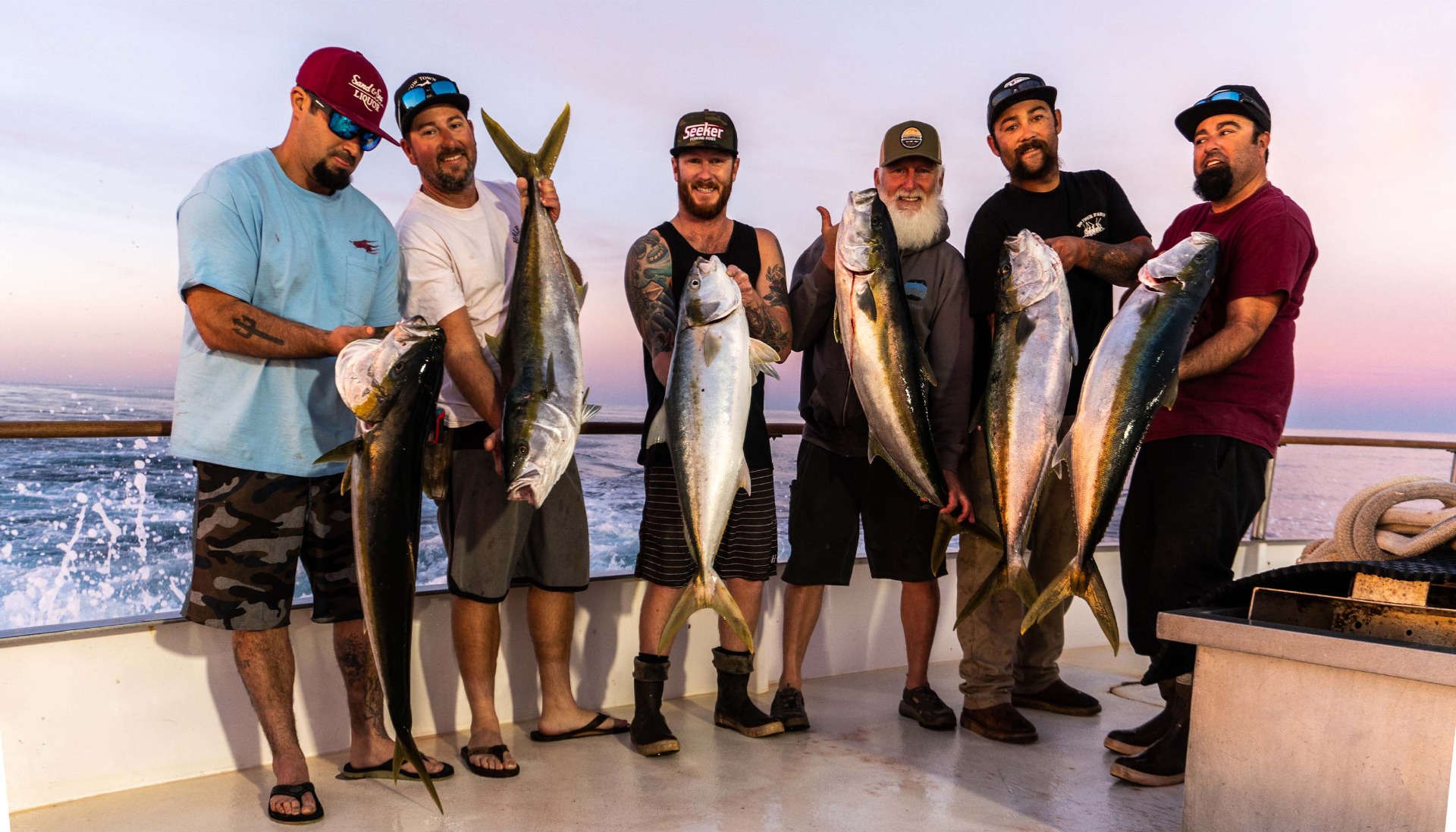 6 Shogun Crew standing on a boat deck at sunset, each holding a large yellowtail fish with silver bodies and yellow tails; most are smiling, wearing casual fishing attire including caps and shorts.