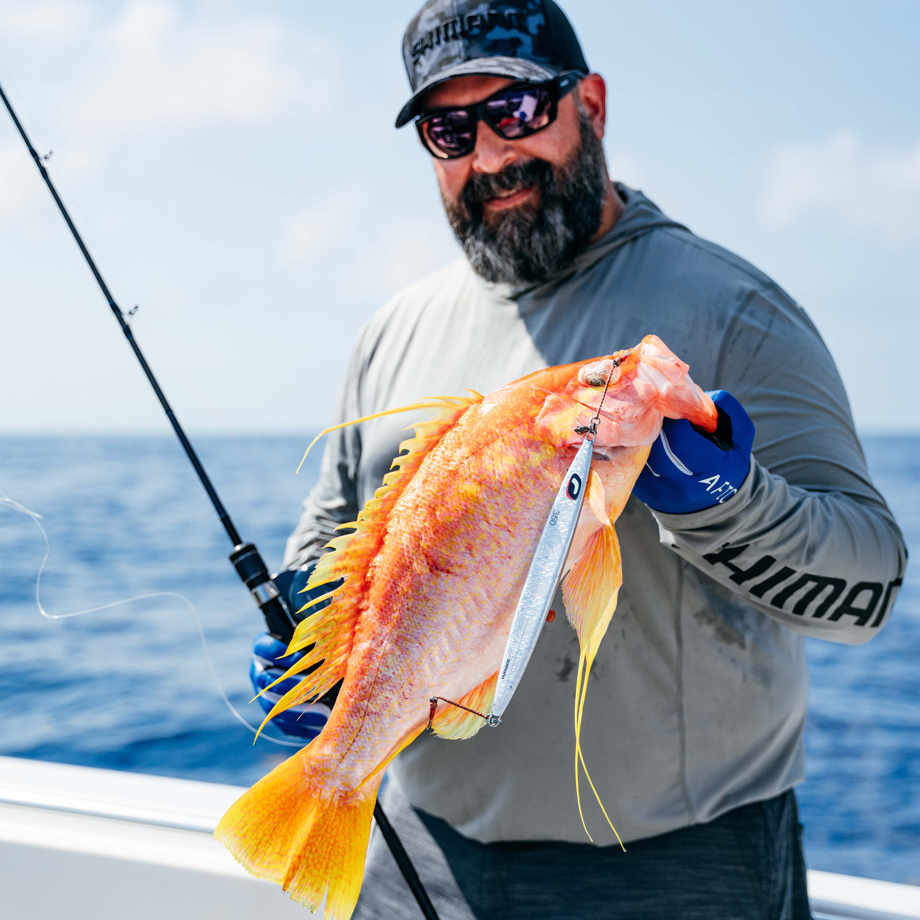 Person on a boat holding a fish with a Shimano Sardine Waver lure attached, while holding a fishing rod over open water.
