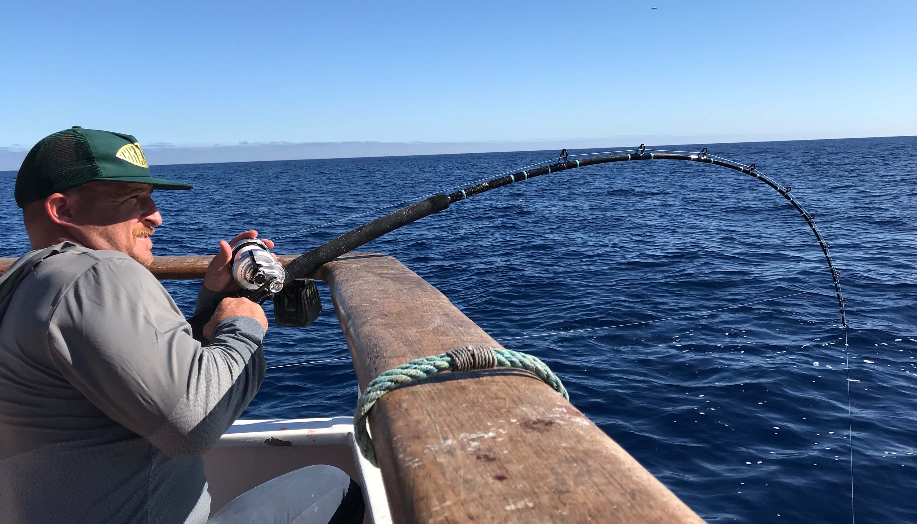 Angler in gray hoodie and green hat bracing at the boat rail, holding a silver lever drag reel with a bent rod under tension. The rod has black and blue wrappings and faces down over open blue ocean beneath a clear sky with horizon visible in background.