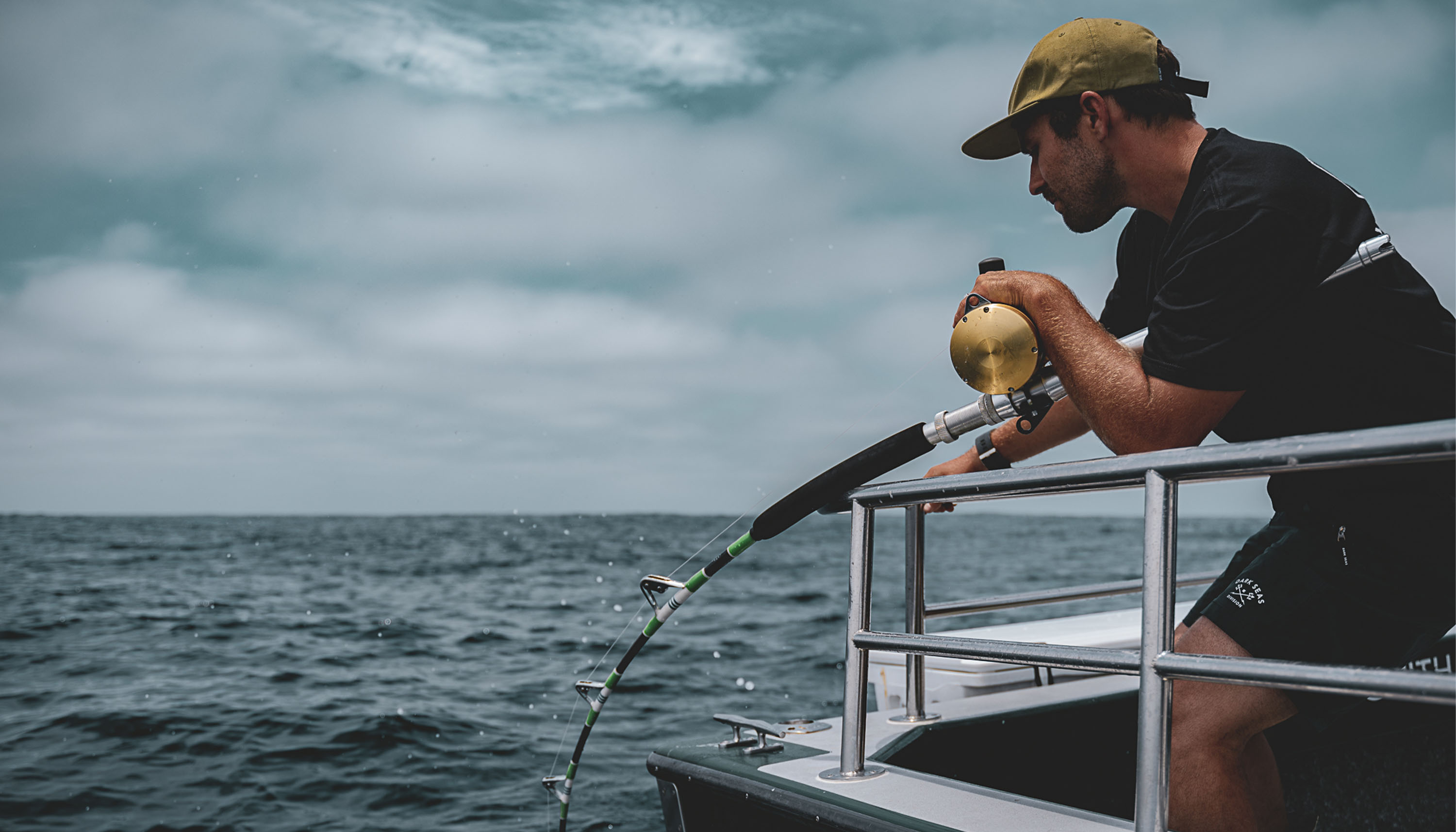 Angler in black shirt and tan cap fishing from a boat rail with a bent rod and gold Tiagra lever drag reel. Rod has green and white wraps with guides facing up. Overcast sky and open ocean fill the background with water splashing near the line.
