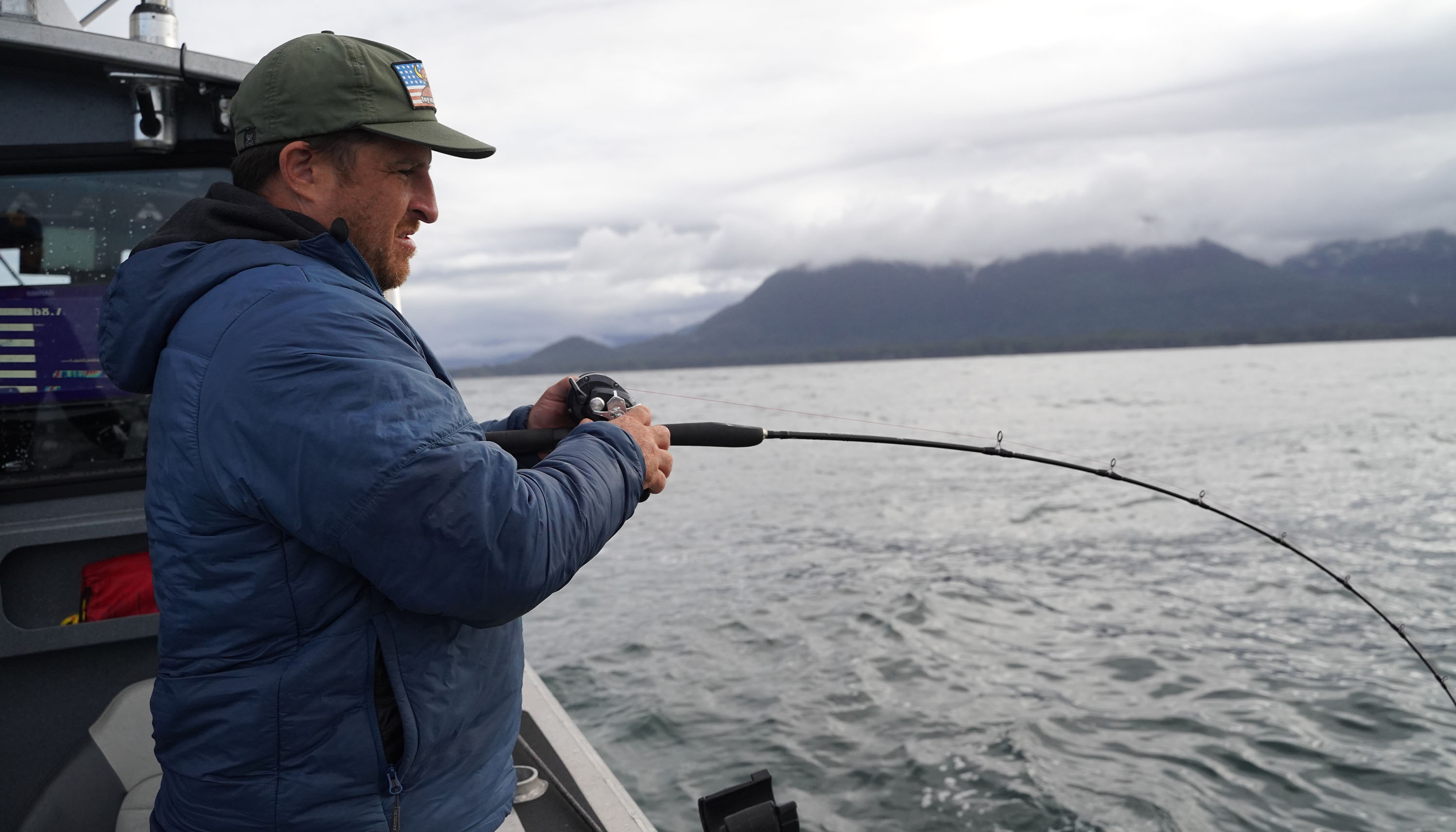 Angler in blue jacket and green cap fishing from a boat using a Torium star drag reel. Rod is bent under tension, with guides facing up. Overcast sky and forested mountains visible across choppy water in the background.