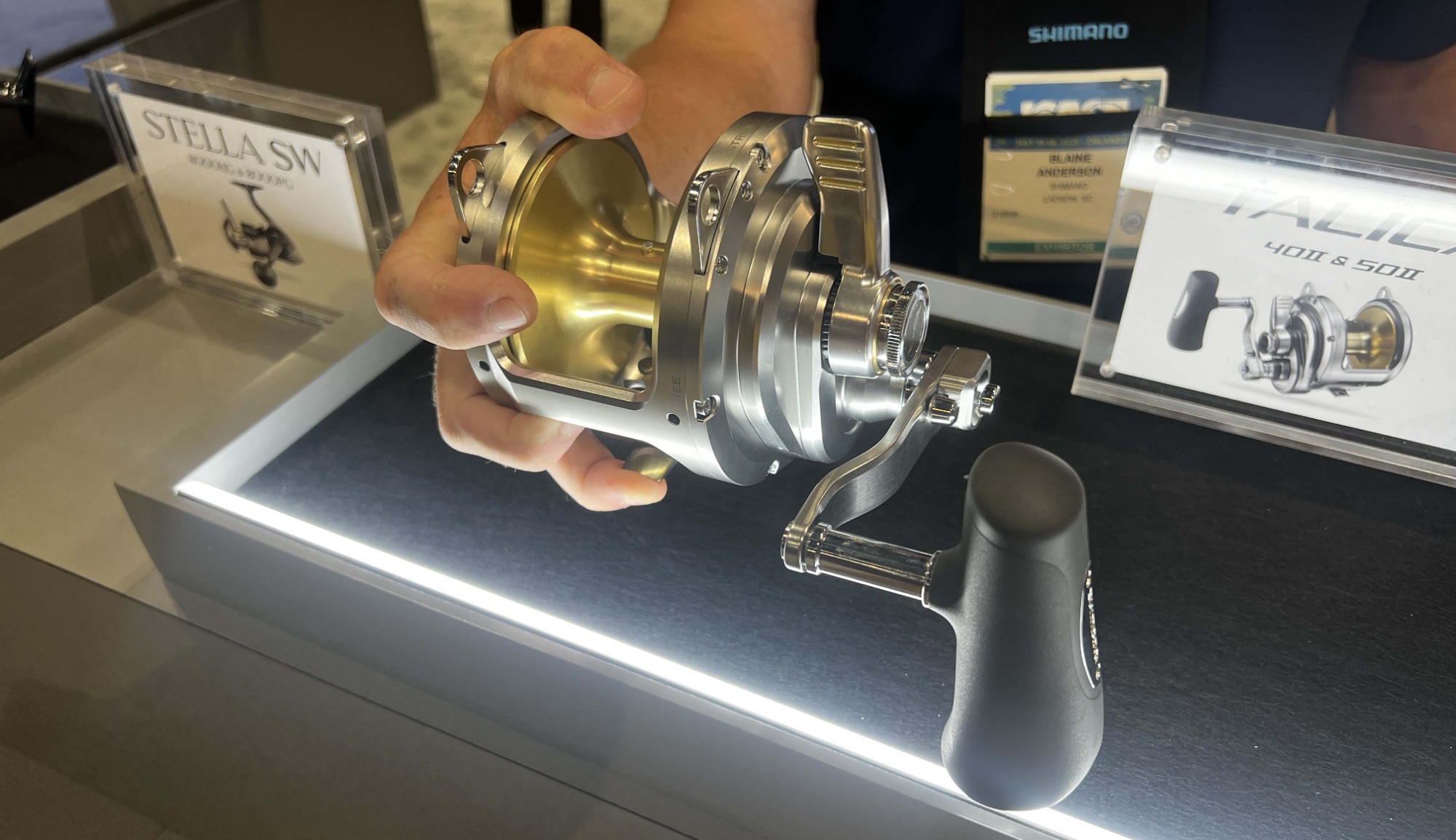 Person holding Shimano Talica 40IIA reel, showing compact size and ergonomic design. Display includes 40II and 50II signage.