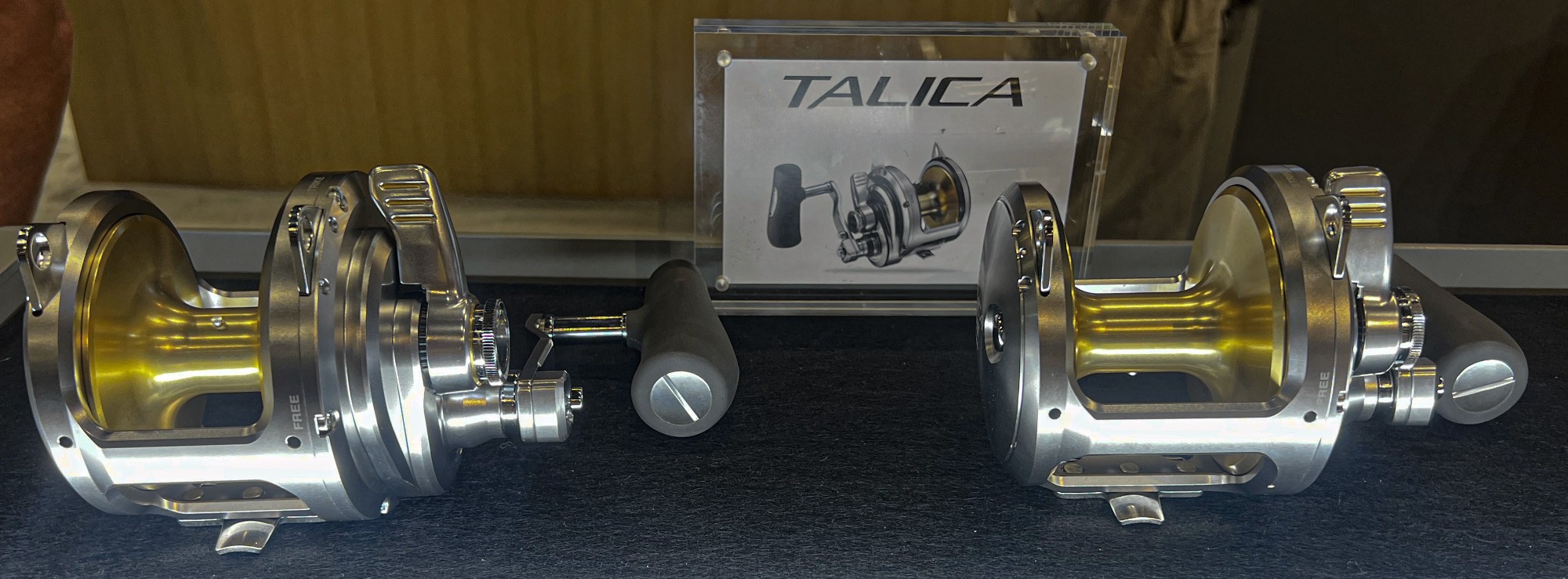 TALICA reels on display with two silver and gold lever drag reels side by side. Background shows a TALICA sign with reel illustration.