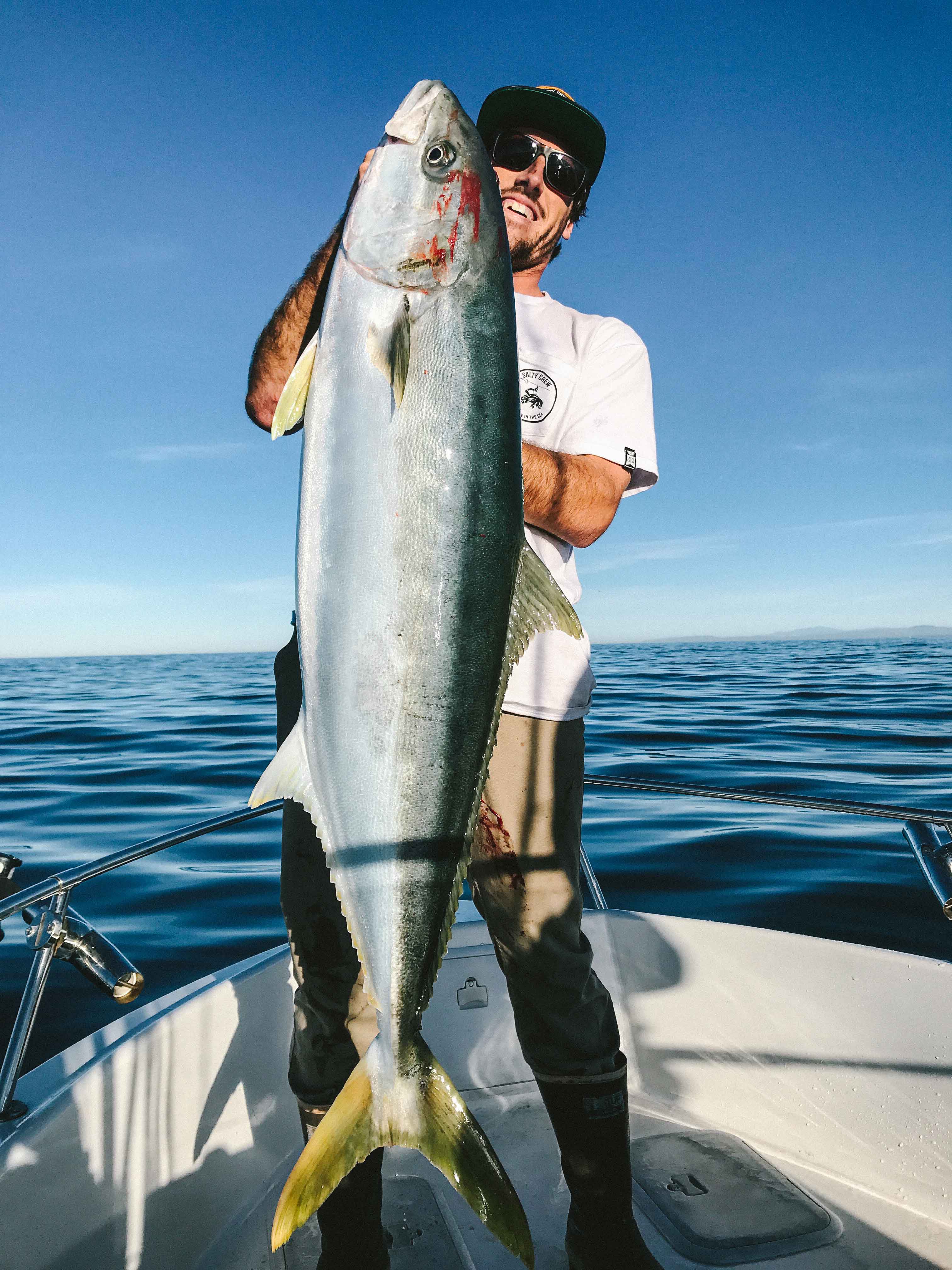 Smiling man in sunglasses and white shirt stands on a boat deck holding a large yellowtail fish vertically. The fish shows blood near the gills. Calm ocean and blue sky in the background.