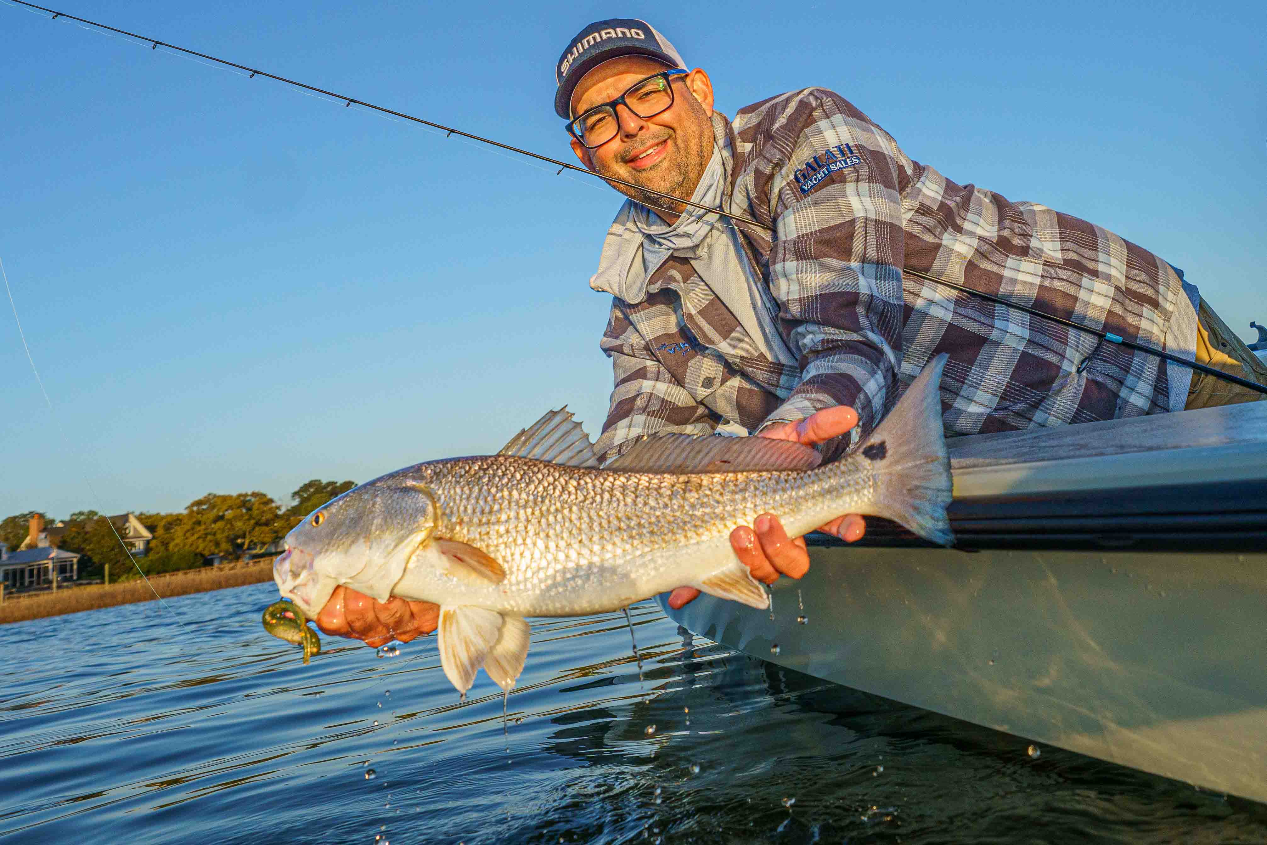 Andrew Alvarez leaning over the side of a skiff at sunrise holding a redfish caught on a green Z-Man soft plastic lure in shallow coastal water.