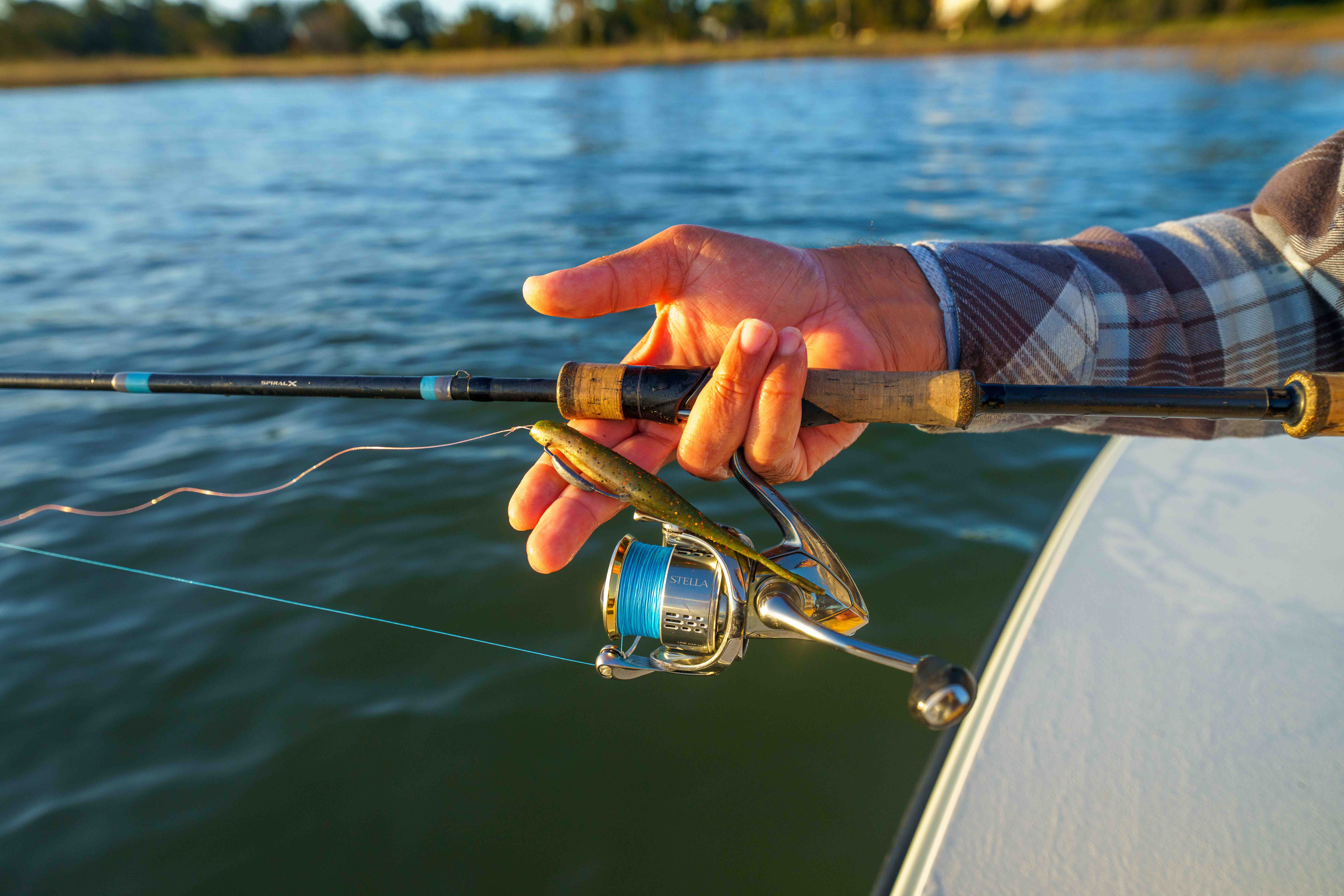 Close-up of an angler holding a spinning rod and reel rigged with a Z-Man soft plastic swimbait over shallow coastal water at sunset.