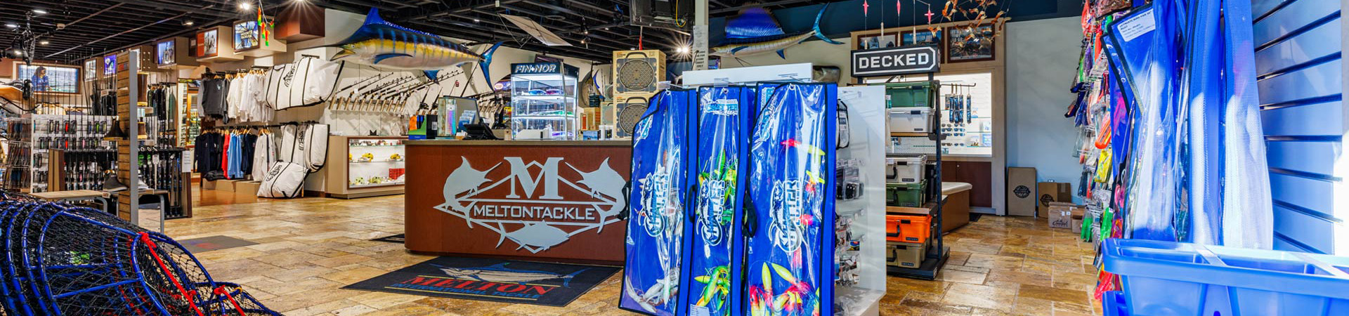 Front desk at Melton Tackle Anaheim retail store with logo on counter and fishing gear throughout. Background shows rods, apparel, lures, and mounted fish displays.